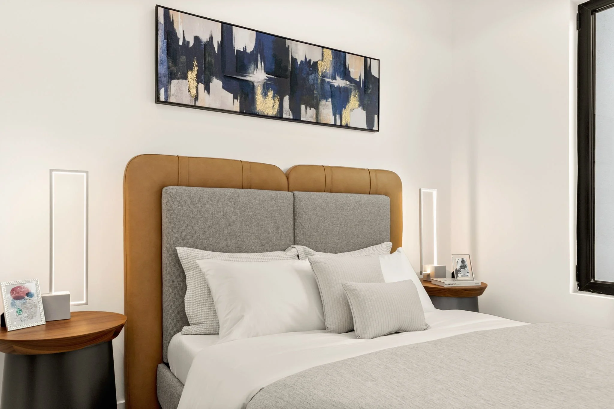 A neatly made bed with white and gray pillows, brown headboard, and side tables in a modern bedroom with white walls, abstract artwork above, and black-framed window.