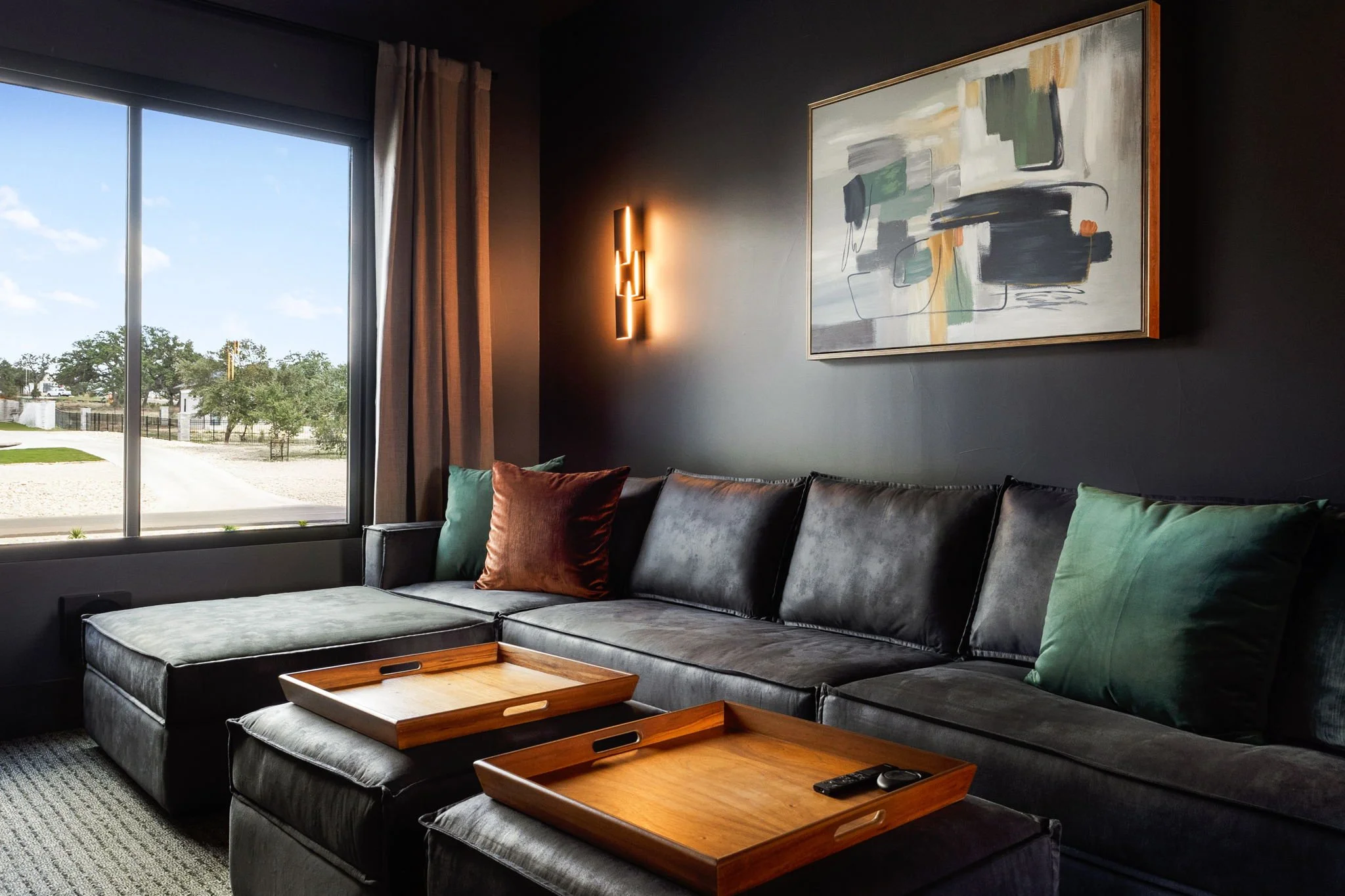 Living room with a large dark sofa, decorative pillows, wooden trays, a window with curtains, an abstract painting, and a wall-mounted light fixture.