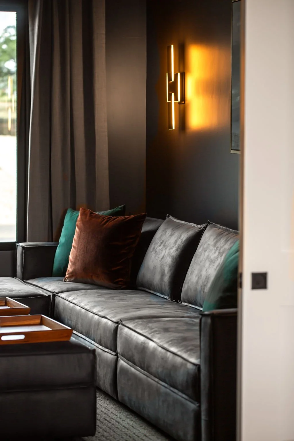 A modern living room with a gray velvet sofa, green and brown throw pillows, a wooden tray on the armrest, dark curtains, and a black wall with a warm yellow illuminated modern wall sconce.