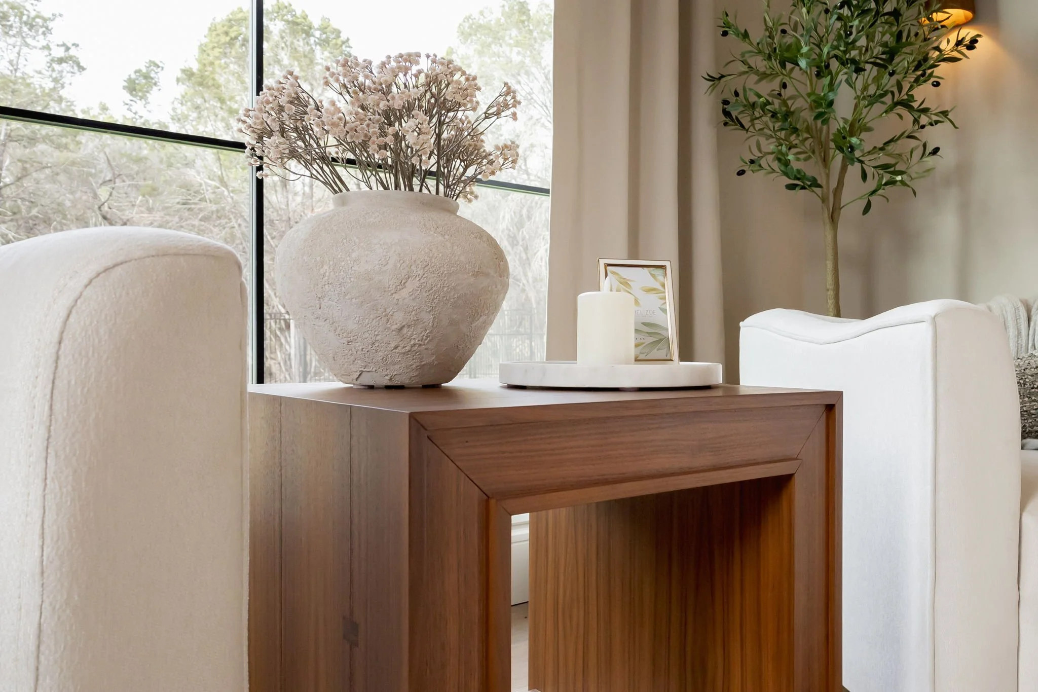 Decorative vases, candle, and picture frame on a wooden table near a window in a cozy living room with white sofa, plants, and neutral curtains
