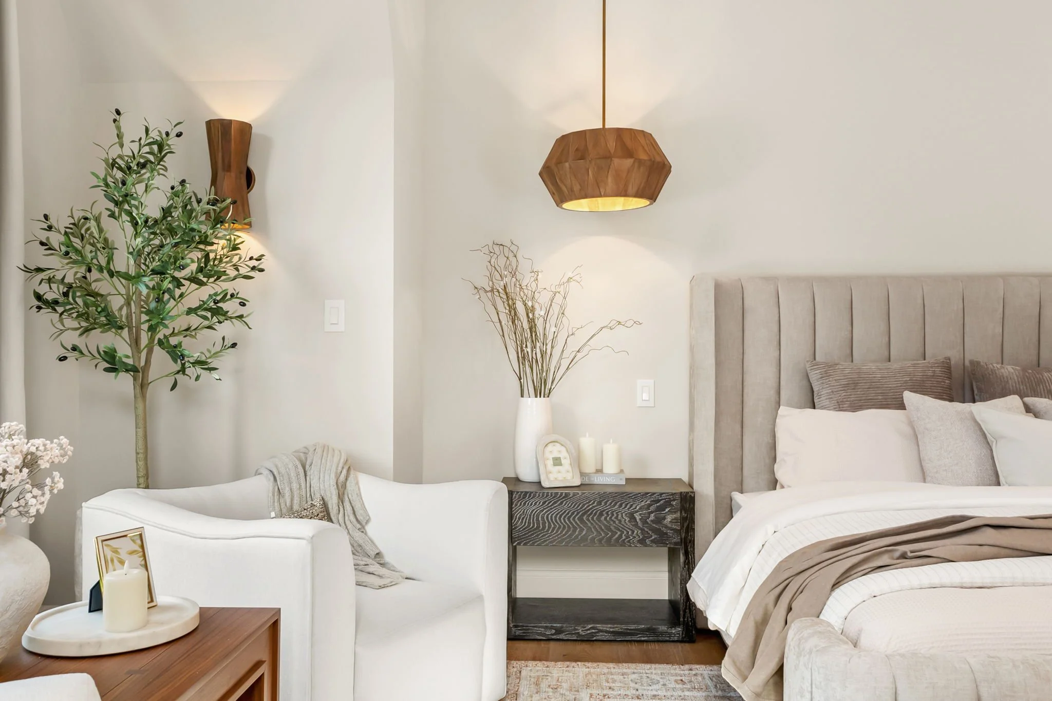 A cozy bedroom features a large upholstered headboard, beige and white bedding, a black side table with candles and decorative items, a white armchair with a throw blanket, a tall potted plant, a wall-mounted wooden light fixture, a pendant lamp, and