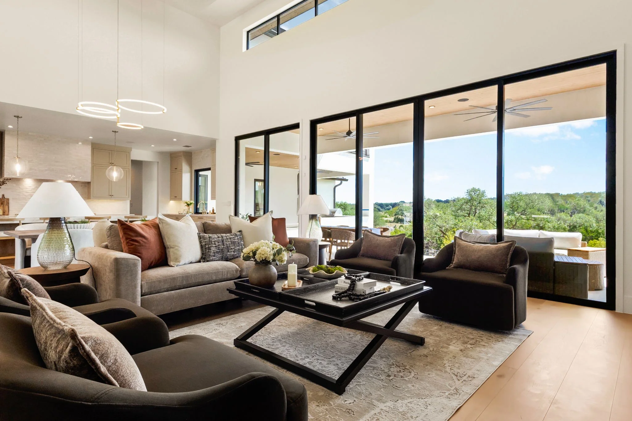 Living room with large glass sliding doors showing green outdoor landscape, modern furniture including sofas and armchairs, coffee table, and decorative items.