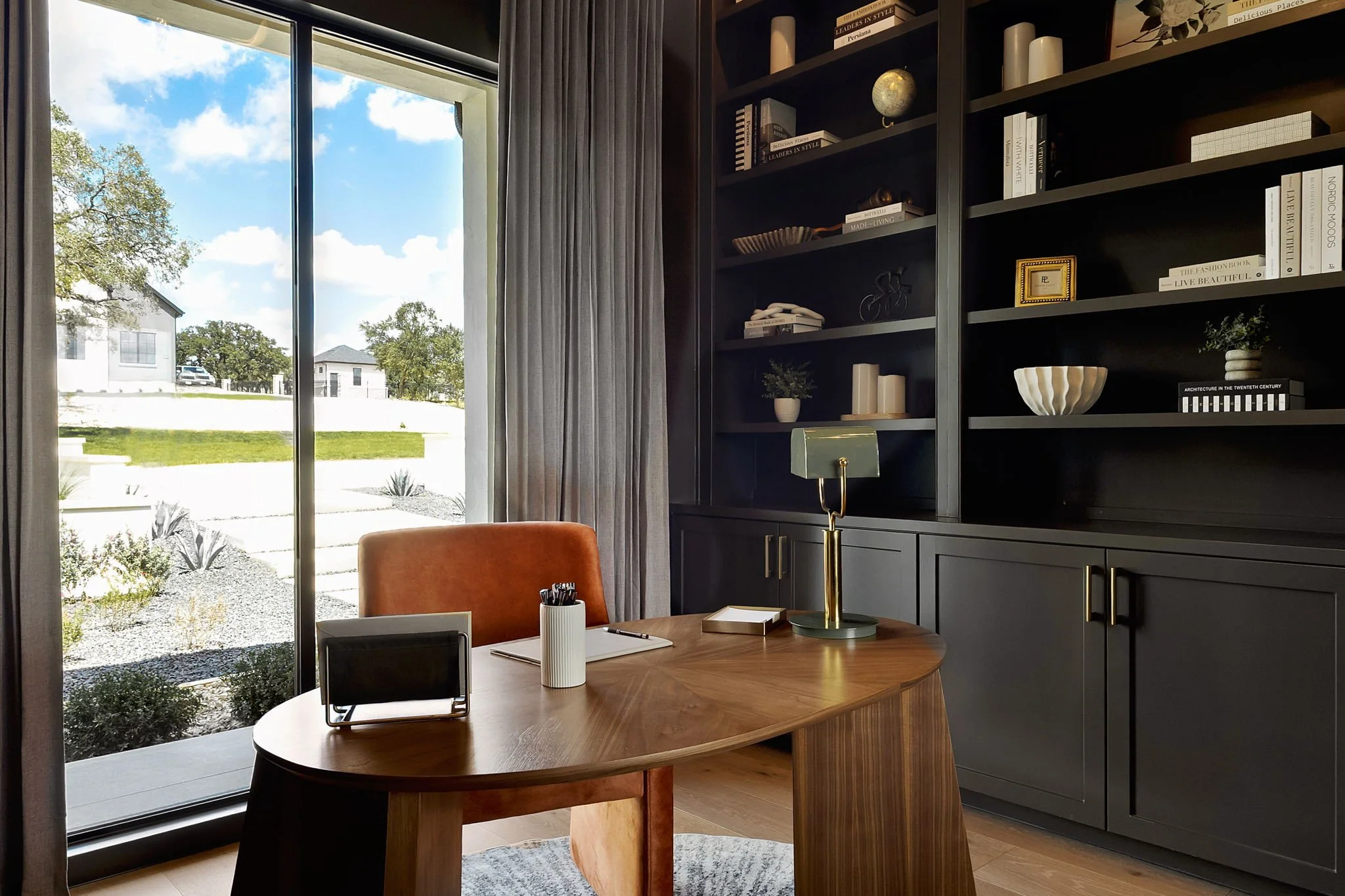Home office with a wooden desk, orange chair, gold desk lamp, tablet, and notepad, facing a large window with gray curtains overlooking a sunny yard with houses and trees.