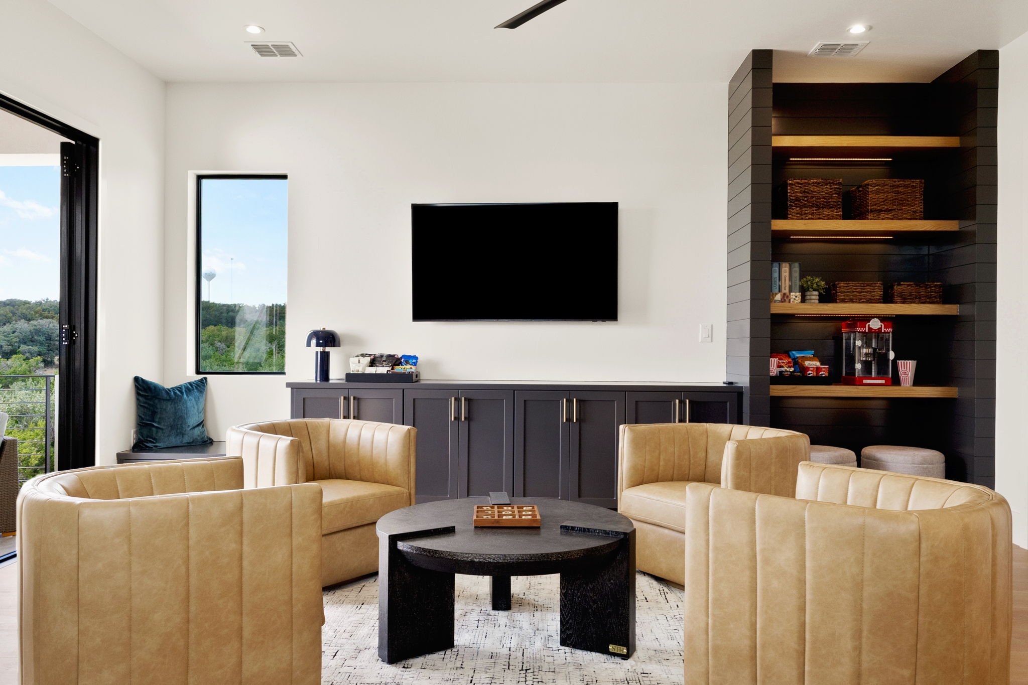 Modern living room with beige leather chairs arranged around a black circular coffee table, white walls, a large flat-screen TV on the wall, a window showing greenery outside, and a built-in shelving unit with baskets and snacks.