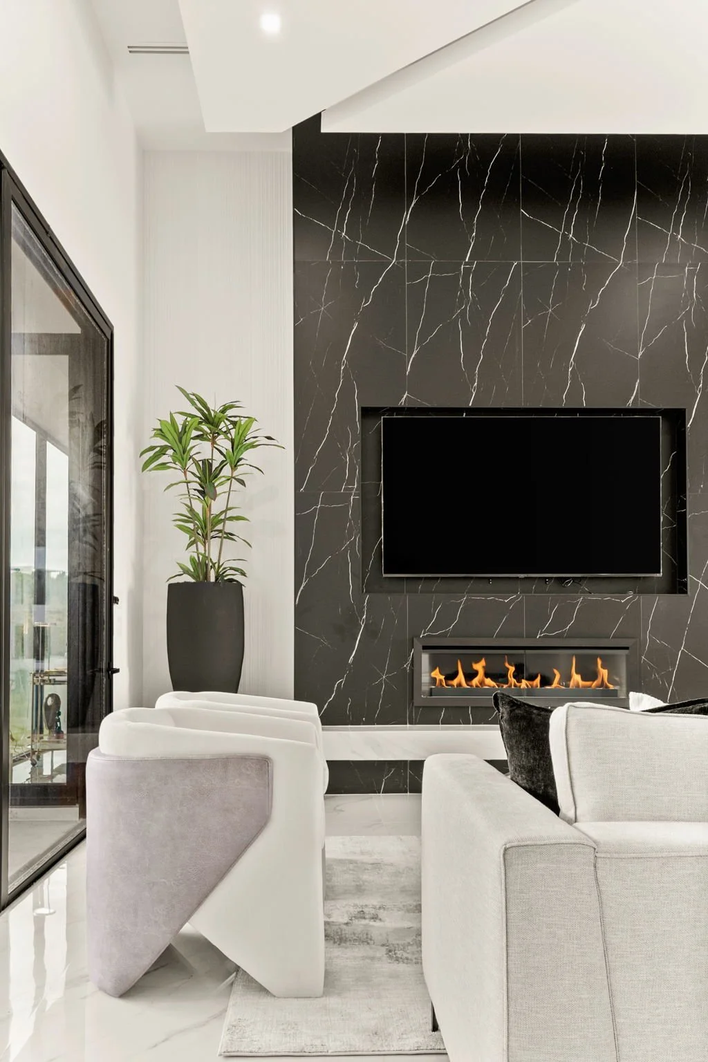 Modern living room with black marble accent wall, flat-screen TV, fireplace with flames, white and gray furniture, black pillow, large potted plant, sliding glass door to outside view.