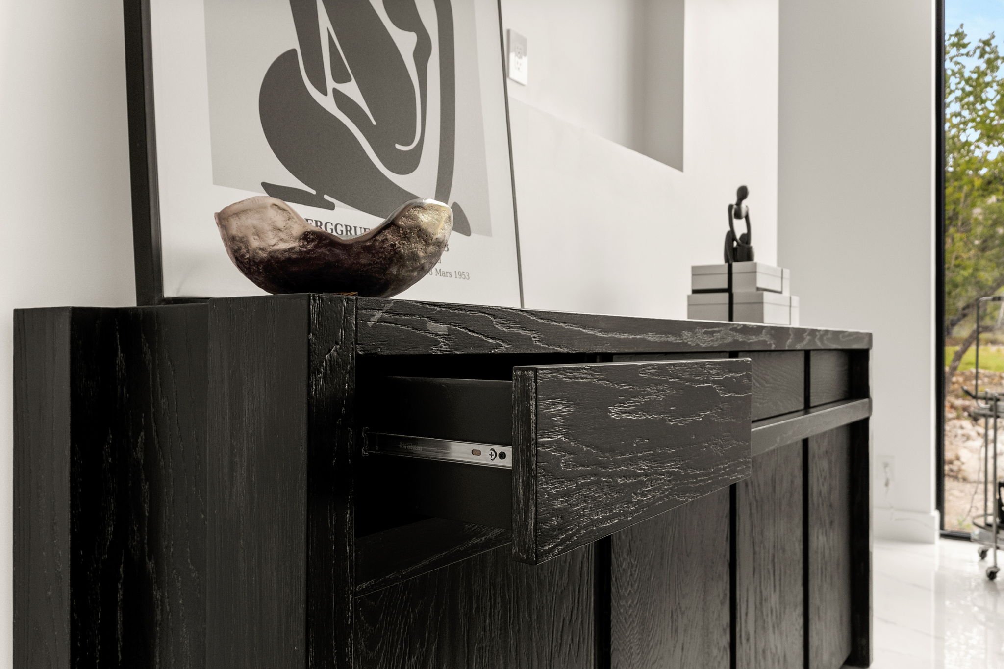 Close-up of a black wooden sideboard with a slightly open drawer, decorated with a ceramic bowl and abstract artwork in the background, in a modern interior setting with natural light.