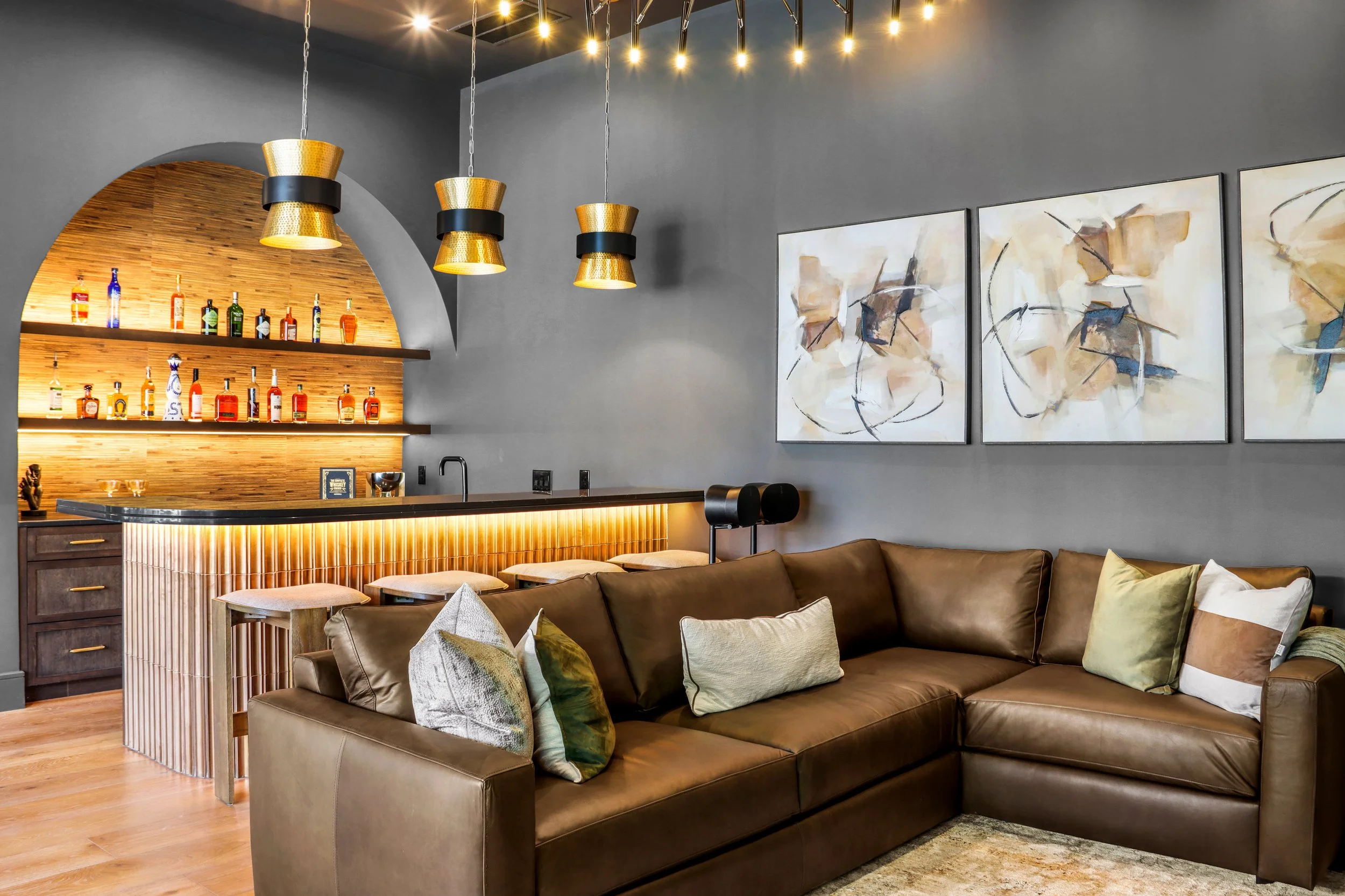 Modern living room with a brown leather sofa, multi-colored throw pillows, abstract wall art, and a bar area with gold pendant lights, wooden accents, and a collection of liquor bottles.