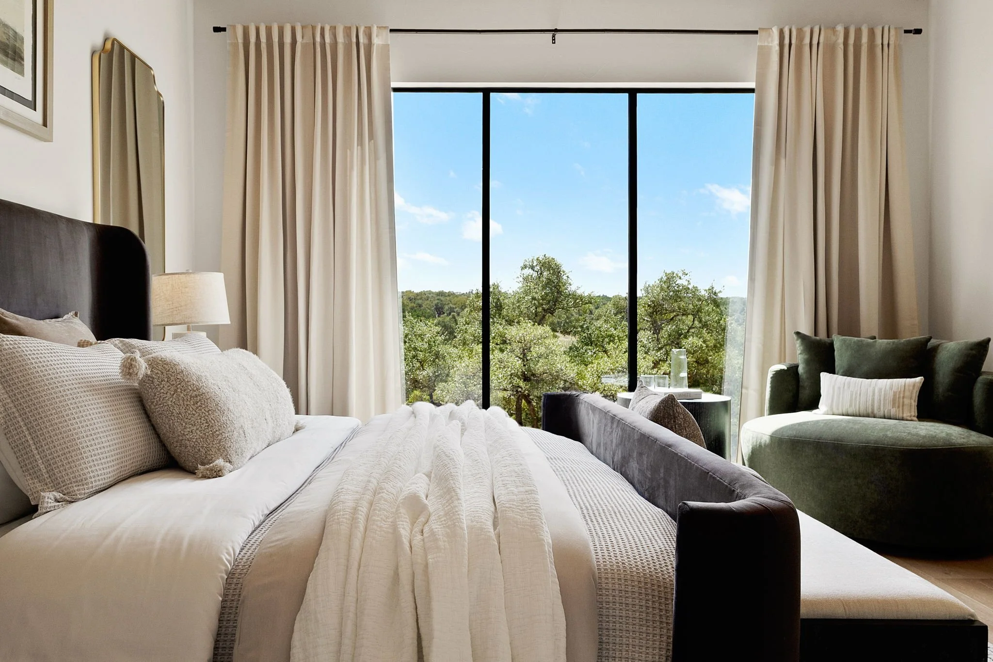 A bedroom with a large bed, cream-colored bedding, and pillows. There are beige curtains, a window showing a view of trees and a blue sky, a green armchair with a pillow, a small side table with a glass of water, and a mirror on the wall.