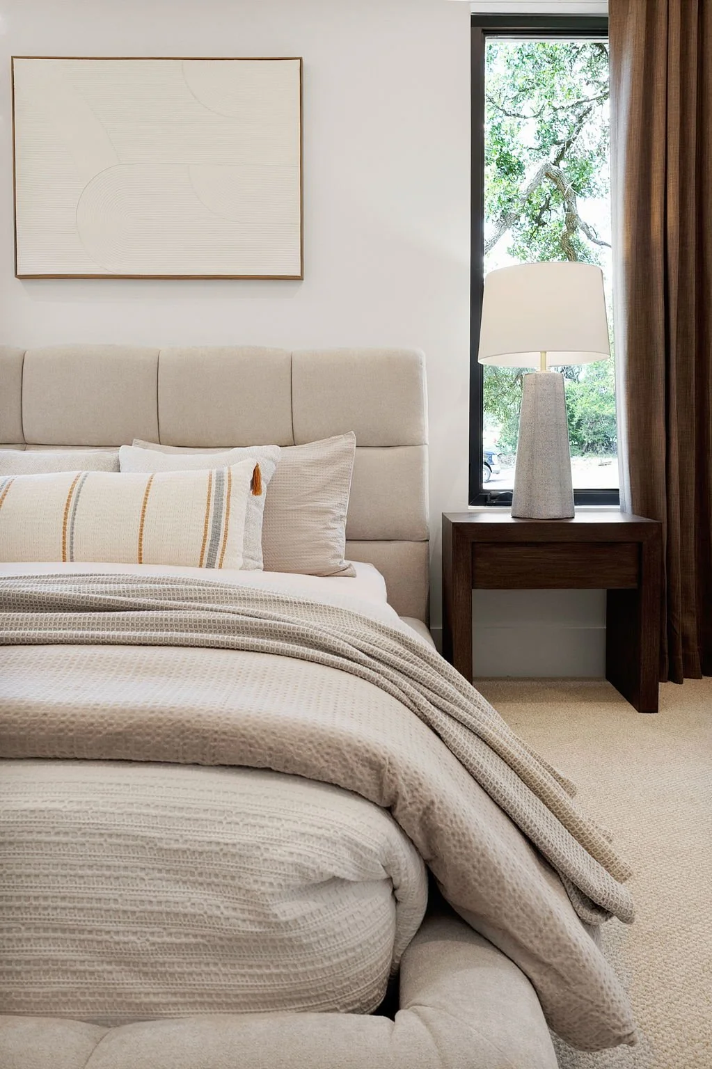 A bedroom with a beige upholstered bed, decorative pillows, a wooden nightstand, a white textured table lamp, and a window showing trees outside.