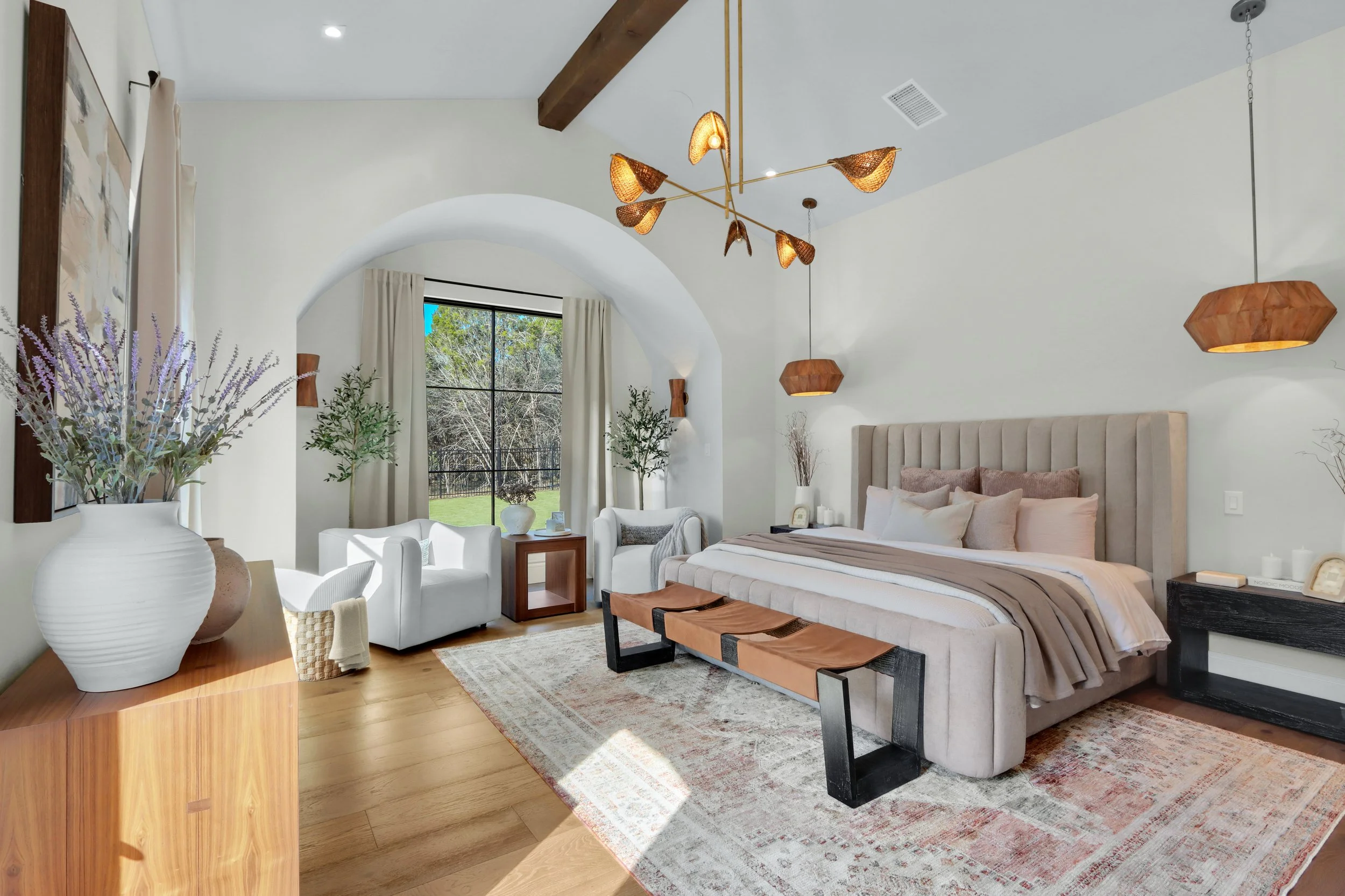 A well-decorated bedroom with a large upholstered bed, hanging pendant lights, and a sitting area near a window with outdoor scenery. There are potted plants, a rug, and modern furniture in neutral tones.