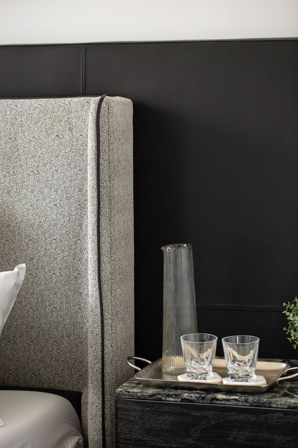 A bedside table with a glass pitcher and two empty glass tumblers on a silver tray, next to a textured beige upholstered headboard and a dark wall.