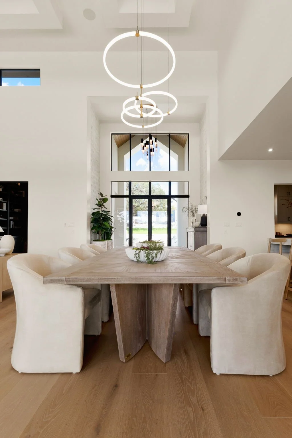 Modern dining room with a large wooden table, surrounded by cream upholstered chairs, and circular pendant lights hanging from the ceiling. Large windows in the background bring in natural light, with a view of a yard and sky outside.