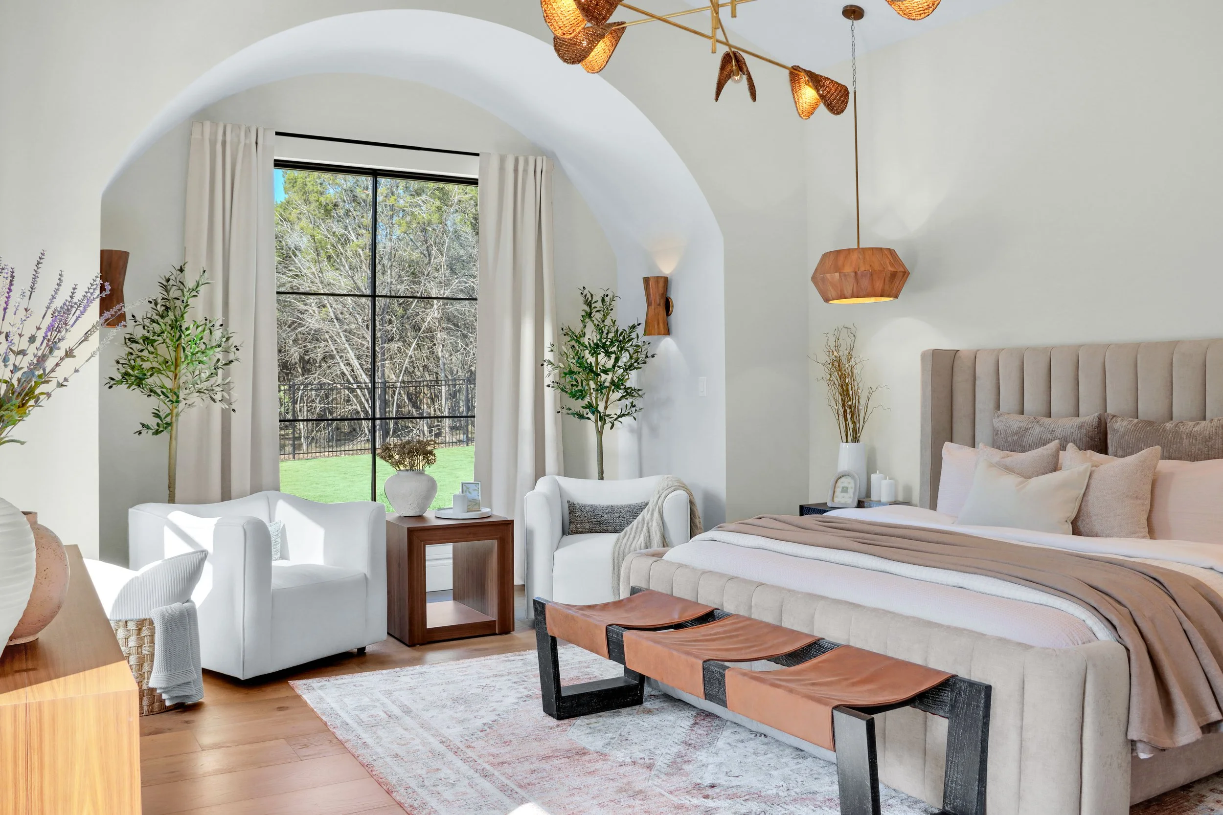 A modern bedroom with a large bed, beige upholstered headboard, multiple pillows, and a blanket. There are two white armchairs, a small wooden side table, and a window with white curtains showing trees outside. The room has wooden flooring, a beige a