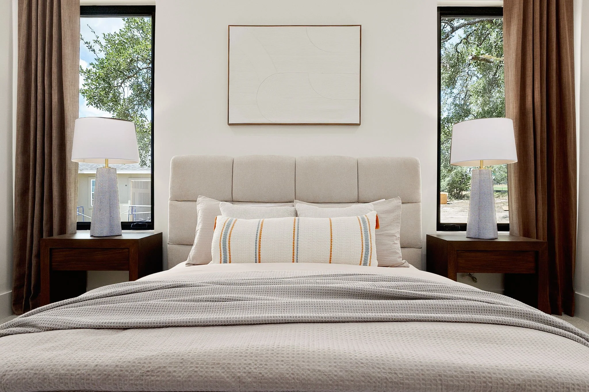 A bedroom with a bed, white headboard, and decorative pillows, flanked by two wooden nightstands with white lamps, windows with brown curtains, and a framed abstract art piece on the wall.