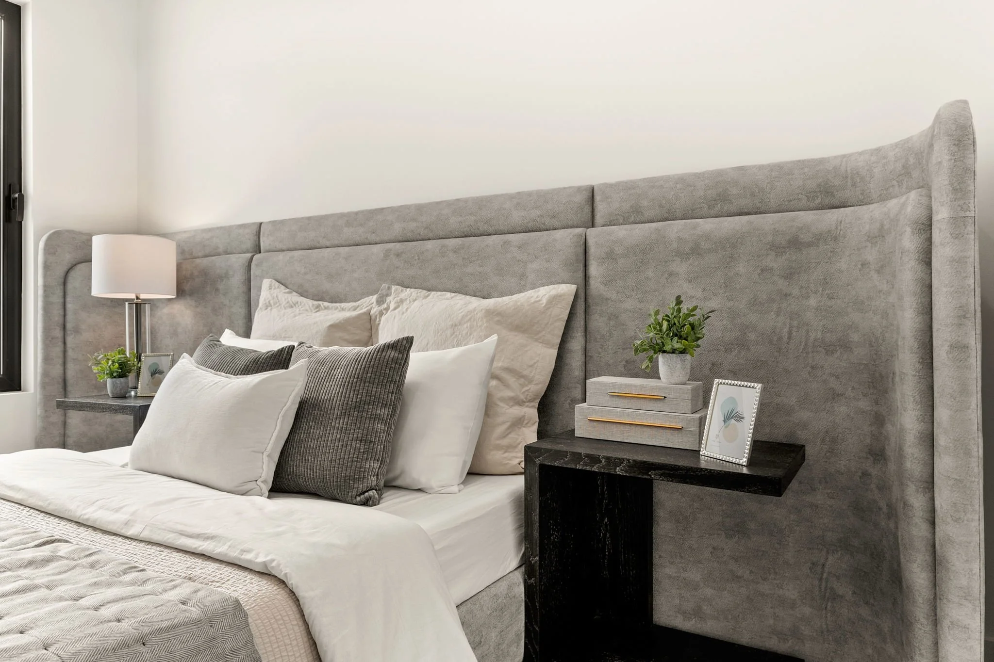 Contemporary bedroom with a large gray upholstered headboard, decorated with multiple white and gray pillows, a bedside black table with a small potted plant, framed art, and stacked books, and a white lamp on a gray wall.