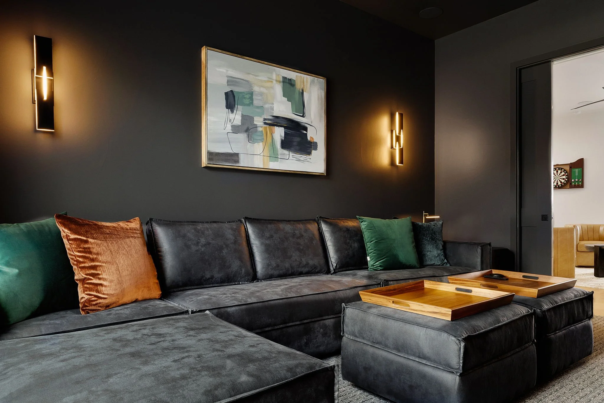 Modern living room with a black leather sectional sofa, colorful pillows, wall art, and wall-mounted lights, with a doorway leading to a room with a dartboard.