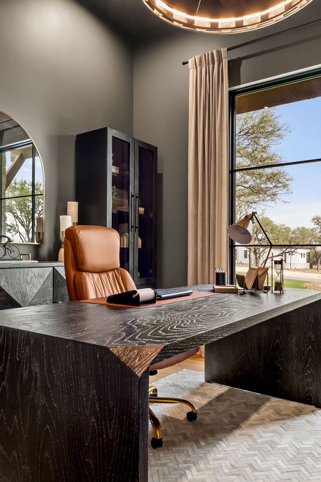 Modern home office with large desk, tan leather chair, black cabinet, large window with beige curtains, and a view of trees outside.