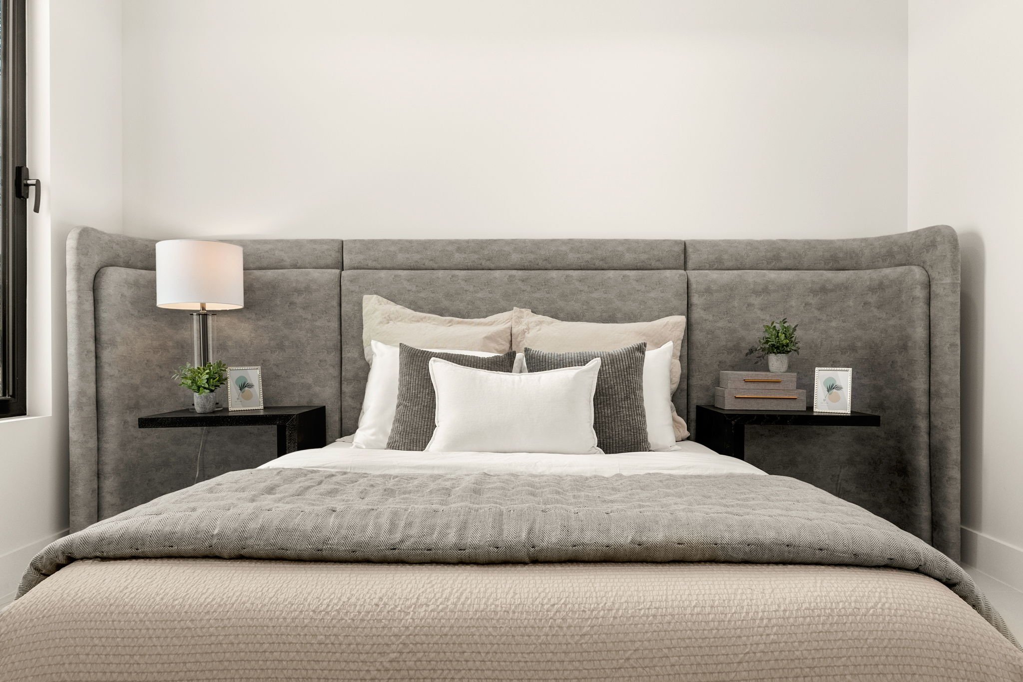 Modern bedroom with a gray upholstered headboard, white walls, and minimalist decor. There are two black nightstands with small plants, framed pictures, and books. The bed has white, gray, and beige bedding with multiple pillows.