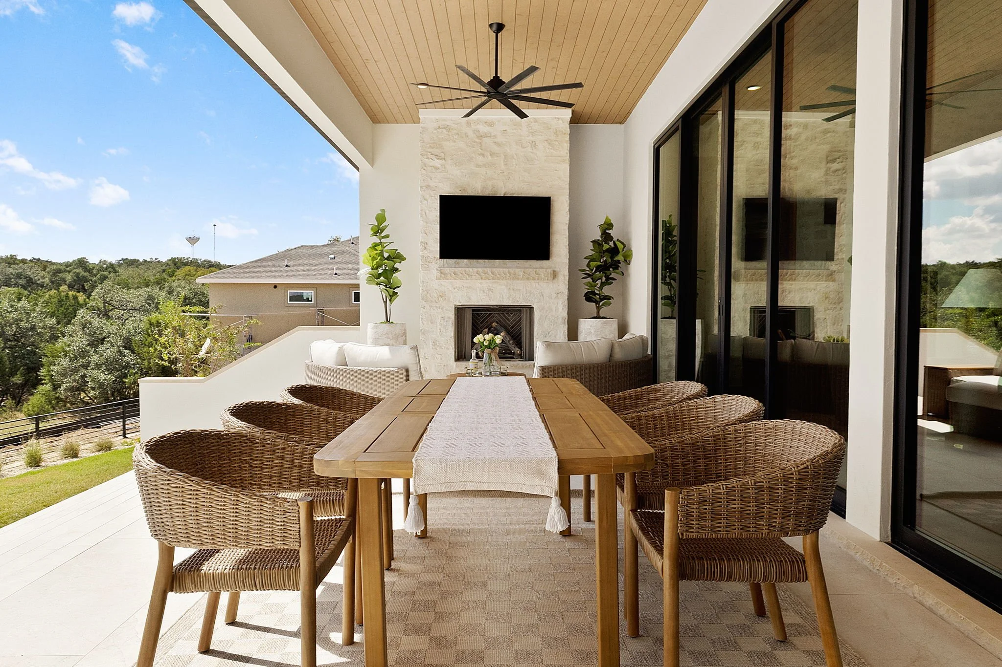Outdoor patio with a wooden dining table, six wicker chairs, a stone fireplace, potted plants, a flat-screen TV, and sliding glass doors.