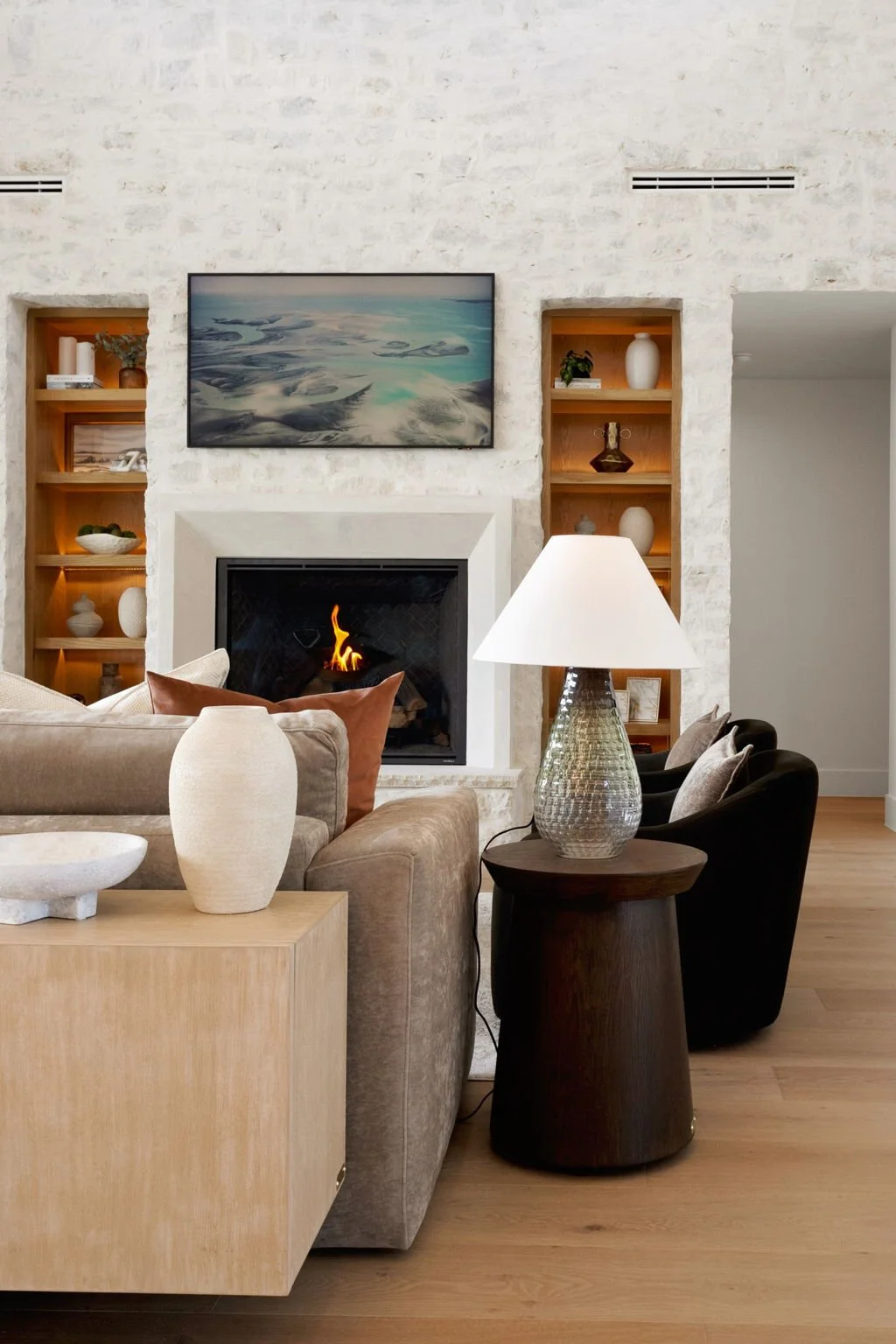Living room with a beige sofa, a tall white vase, a dark side table, a lamp, a black armchair, a fireplace, built-in wooden shelves, a wall-mounted TV, and decorative objects.