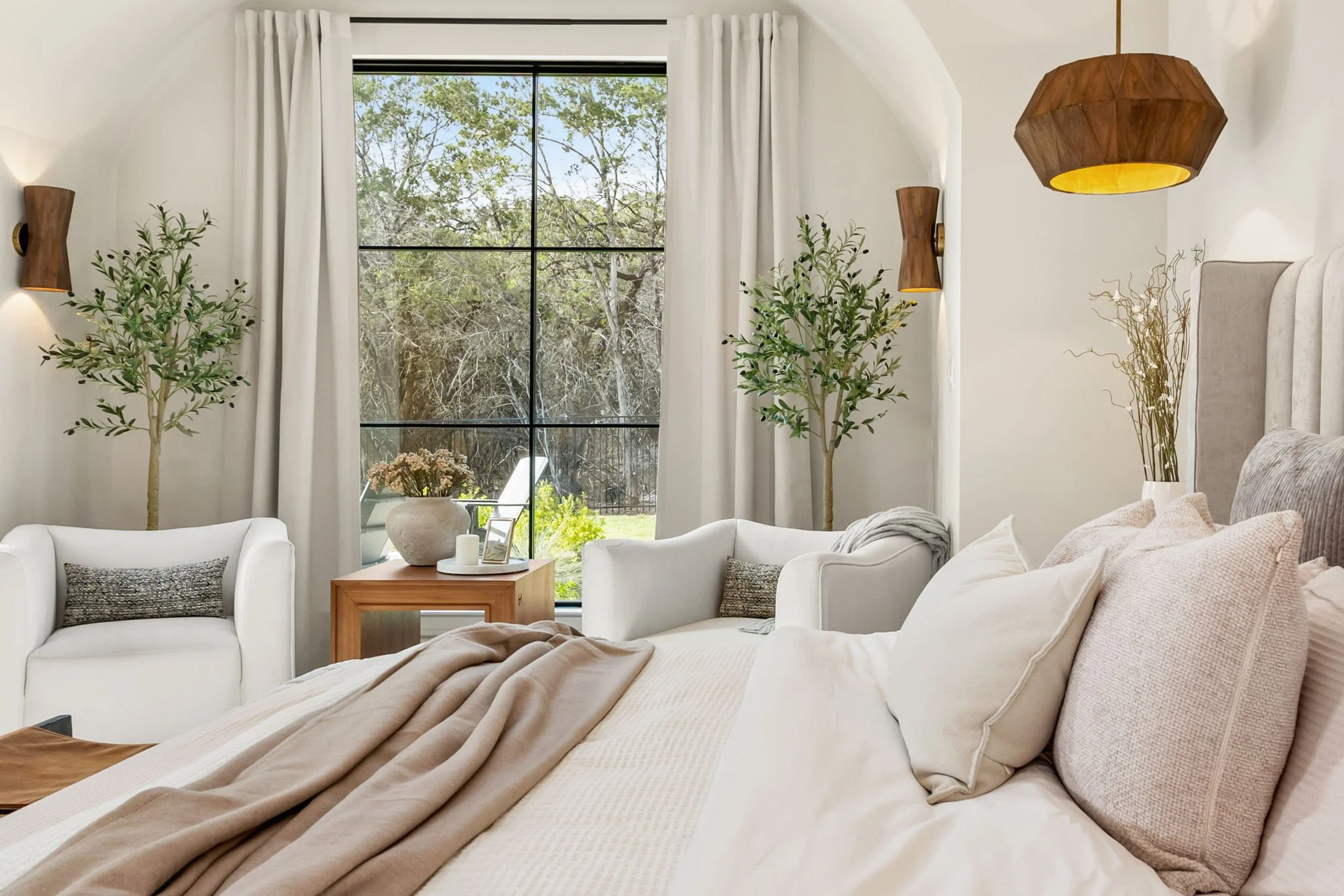 Bright bedroom with a large window showing trees outside. There are two white armchairs, a small wooden side table with a potted plant, and a bed with beige and white bedding and pillows. The room has white curtains, wooden wall sconces, and a ceilin