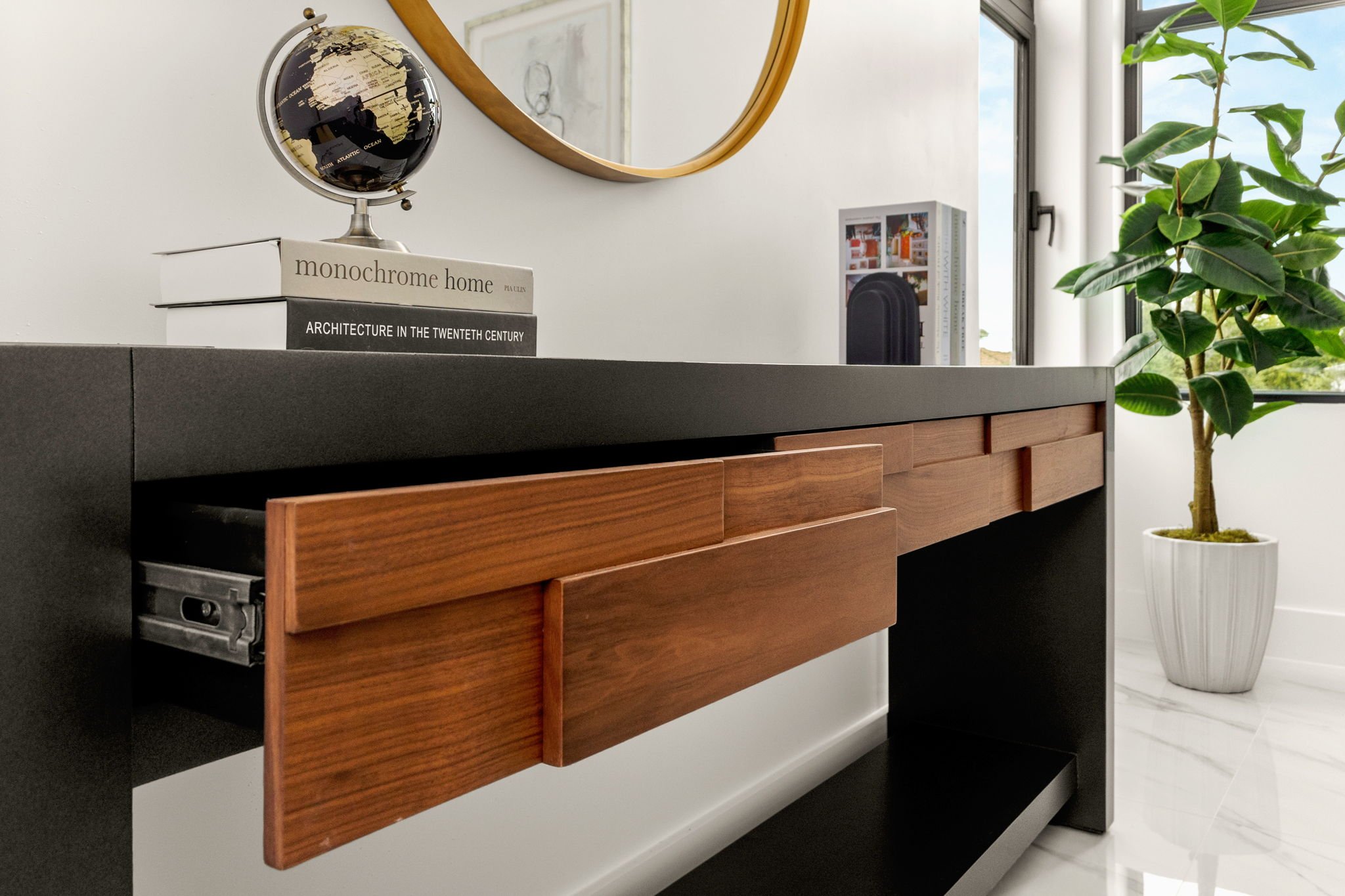 Close-up of a modern living room black and wooden sideboard with books, a globe, and a round mirror above it, next to a large window with a potted plant.