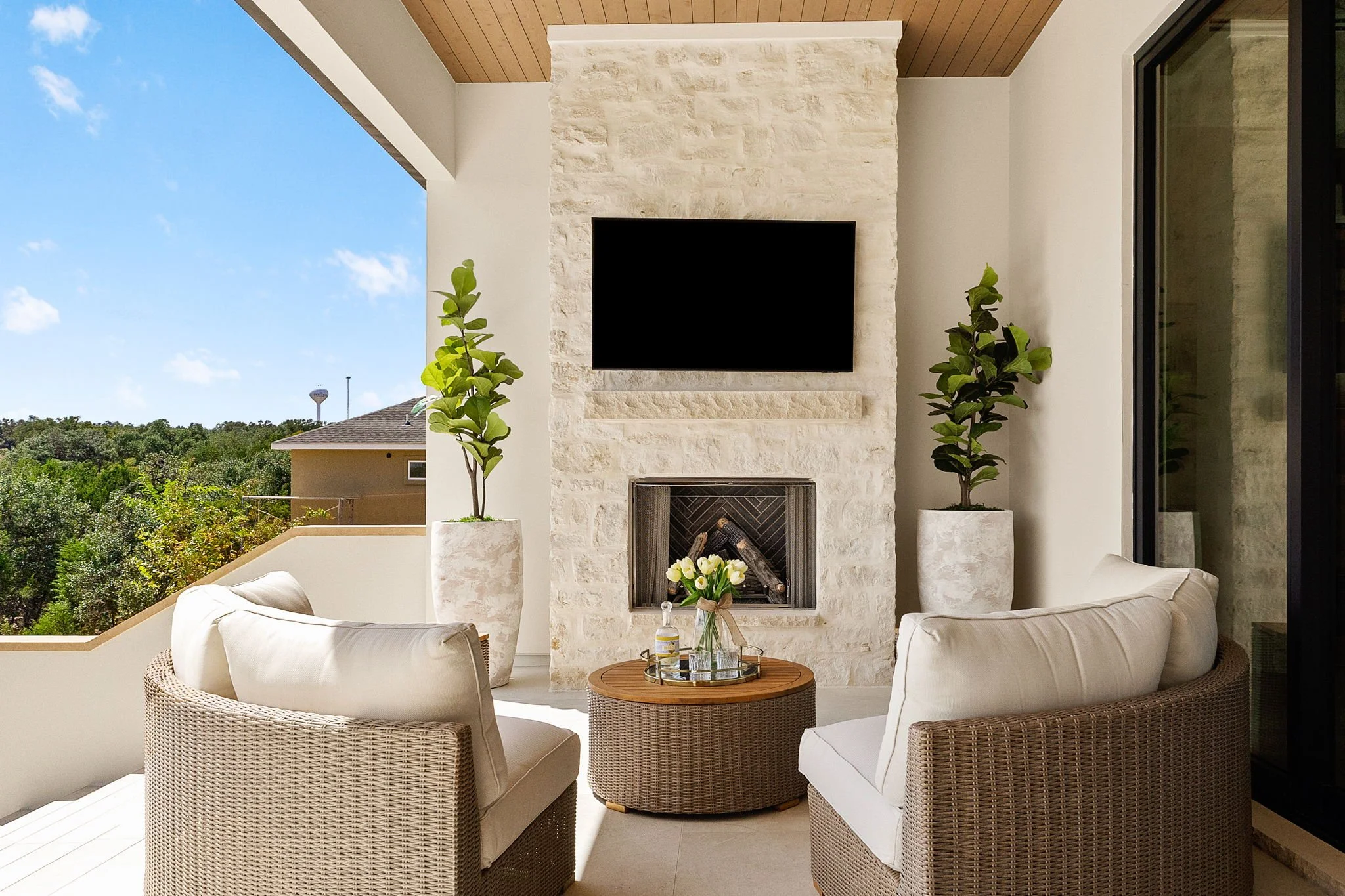 Outdoor patio with beige wicker chairs, a gnome, and a small round table with a water bottle, glasses, and a floral arrangement, facing a stone fireplace with a mounted black flat-screen TV, flanked by two tall potted plants, with a partial view of a