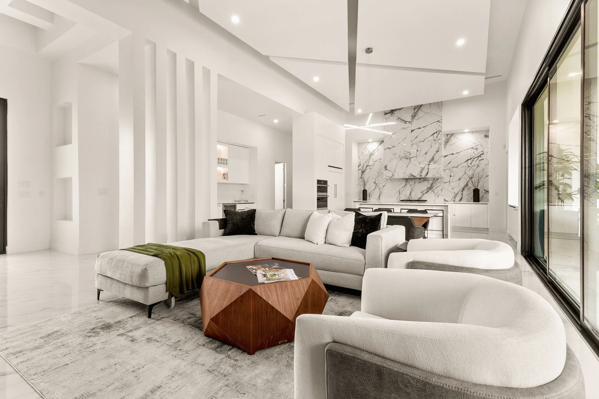 Modern living room with white walls, large window, and marble accent wall, featuring a sectional sofa with white and black cushions, a geometric wooden coffee table, and a patterned area rug.