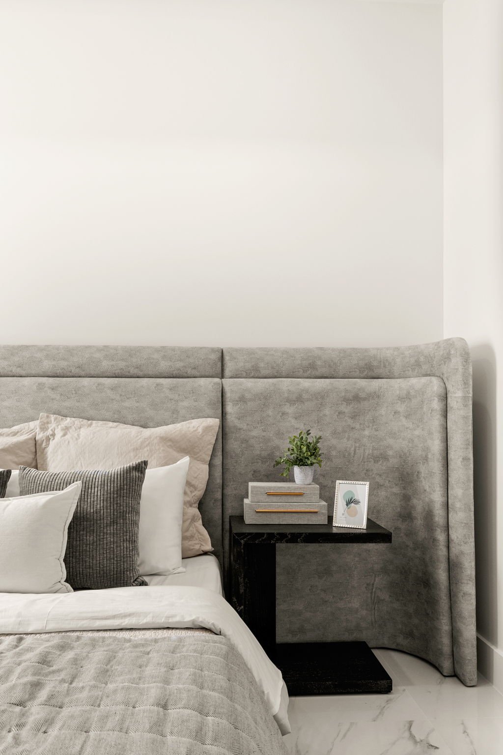 A bedroom with a gray upholstered bed and a black nightstand. The nightstand has a green potted plant, some stacked books, and a framed art print. The wall behind is plain white, and part of the marble floor is visible.