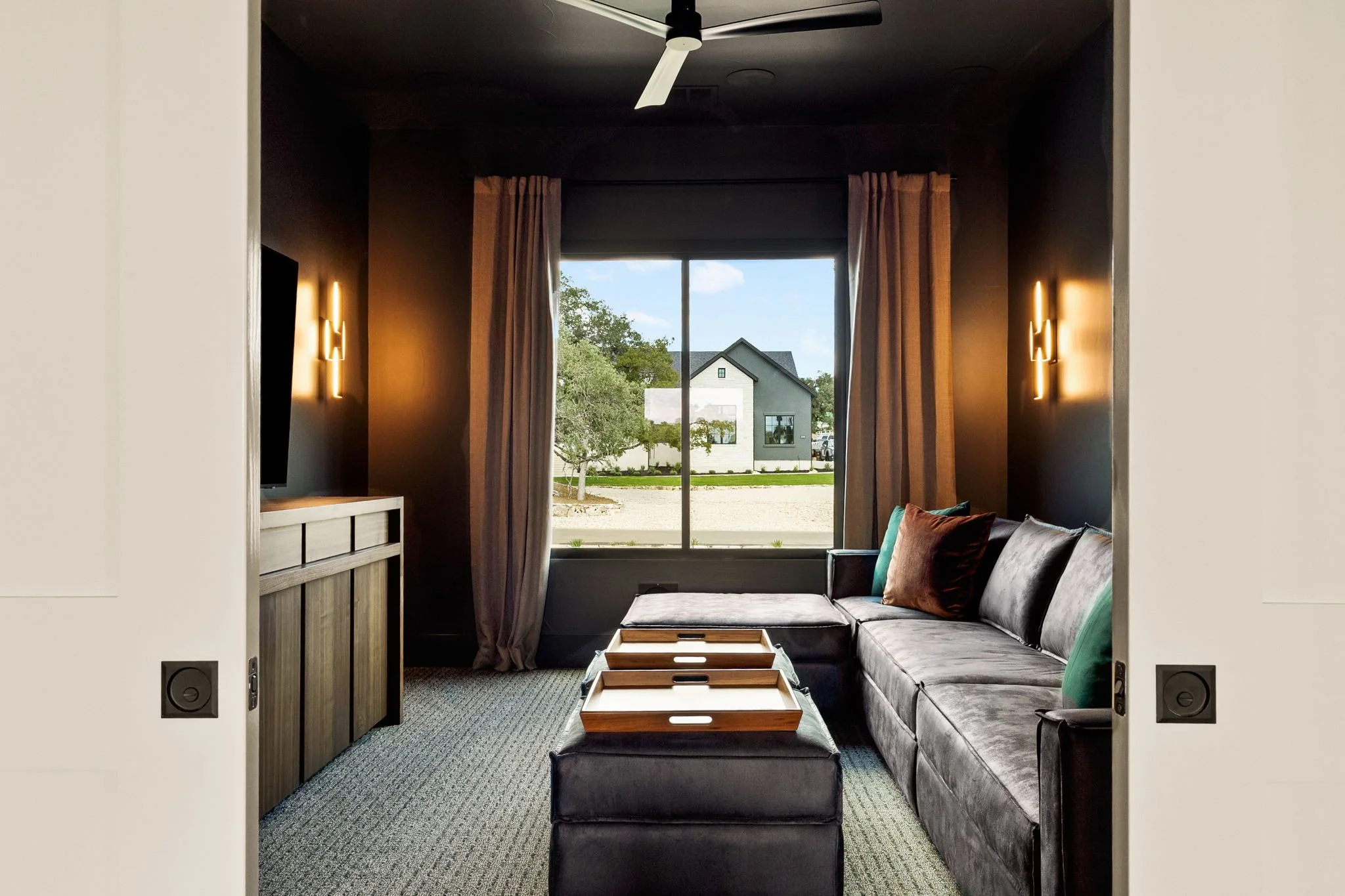 Living room with a large window, gray sectional sofa, throw pillows, wooden trays on an ottoman, dark accent walls, wall sconces, and a ceiling fan.
