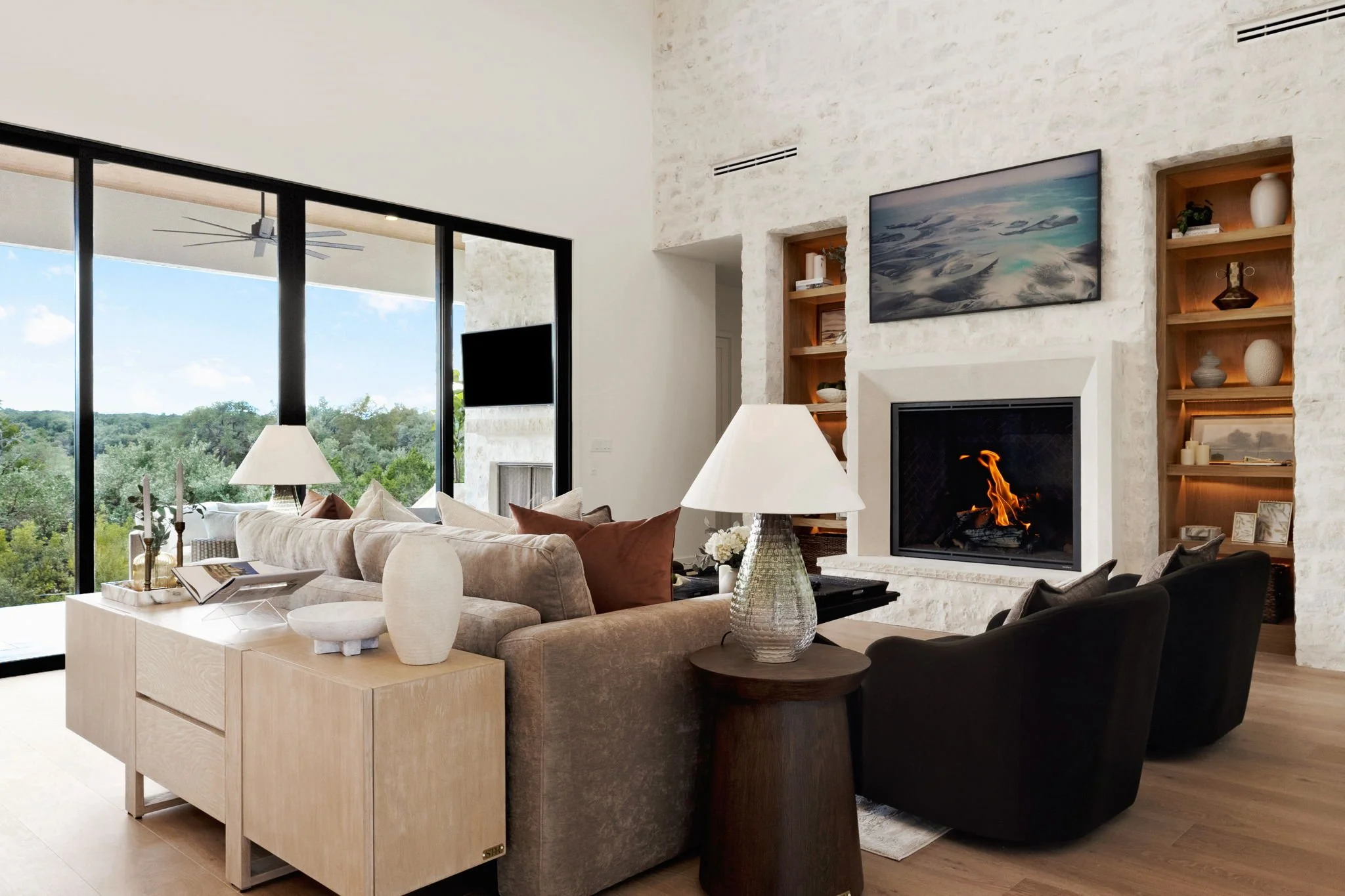 Modern living room with a beige sectional sofa, black armchair, and wooden side table and lamps. A fireplace with a mounted TV above and built-in shelves on the side, with large glass windows showing green trees and blue sky outside.