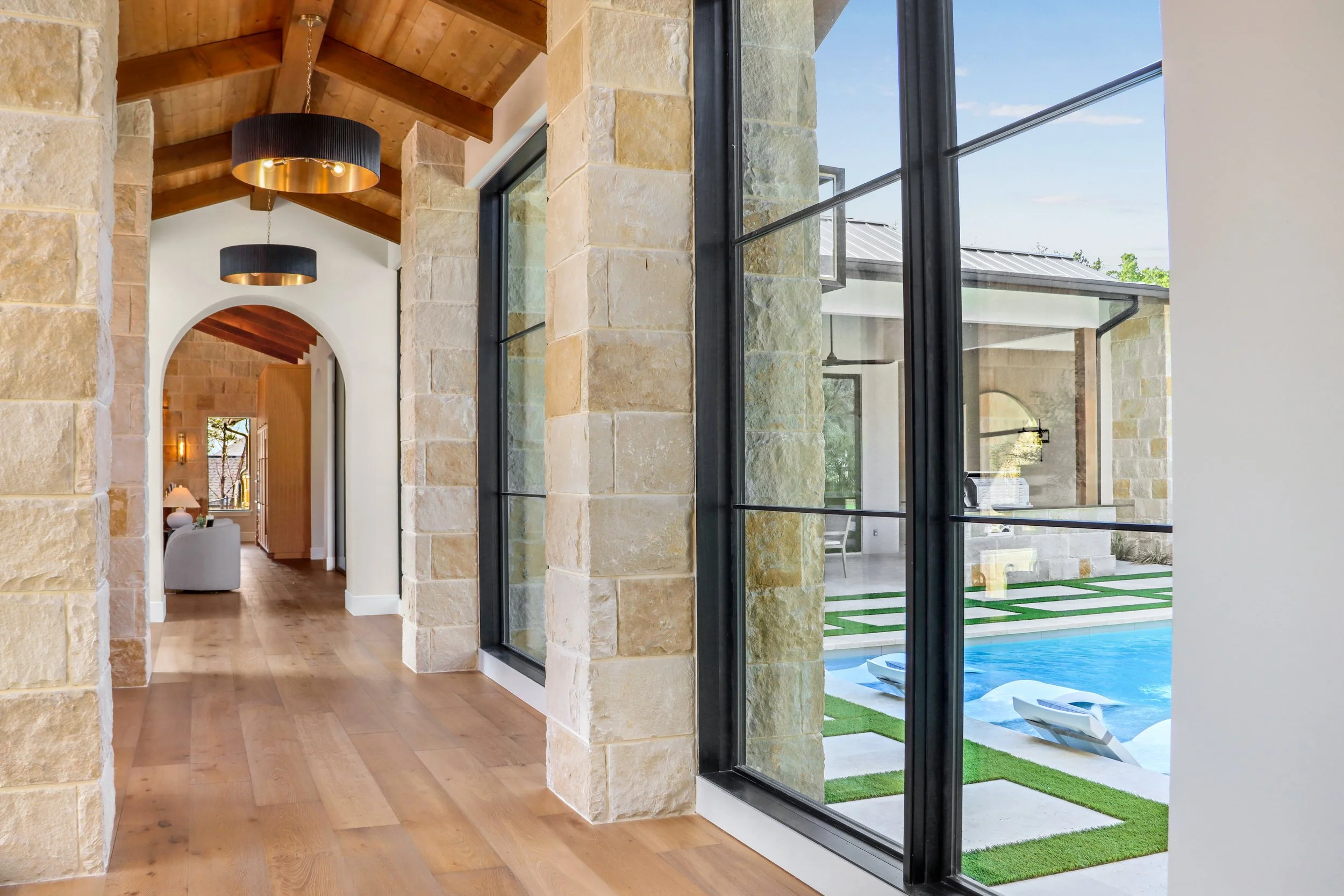 Interior hallway with wood flooring, stone walls, vaulted wooden ceiling, and large windows looking outside at a swimming pool.