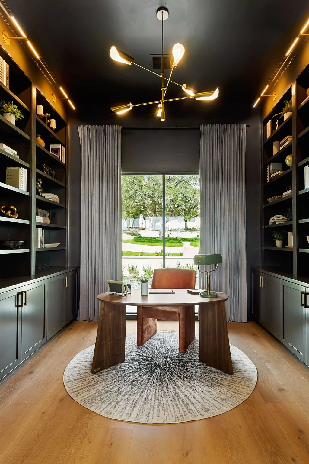 An elegant home office with dark walls, large window with gray curtains, a wooden desk, and a modern chandelier.