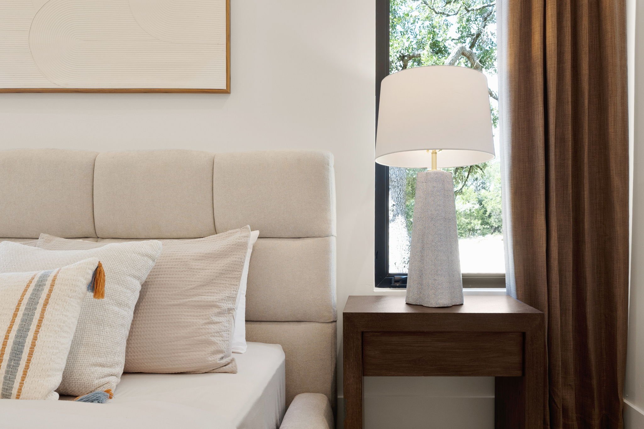 A bedroom with a beige upholstered headboard, beige pillows with tassels, a wooden nightstand, a white table lamp, a window with a view of trees, and brown curtains.