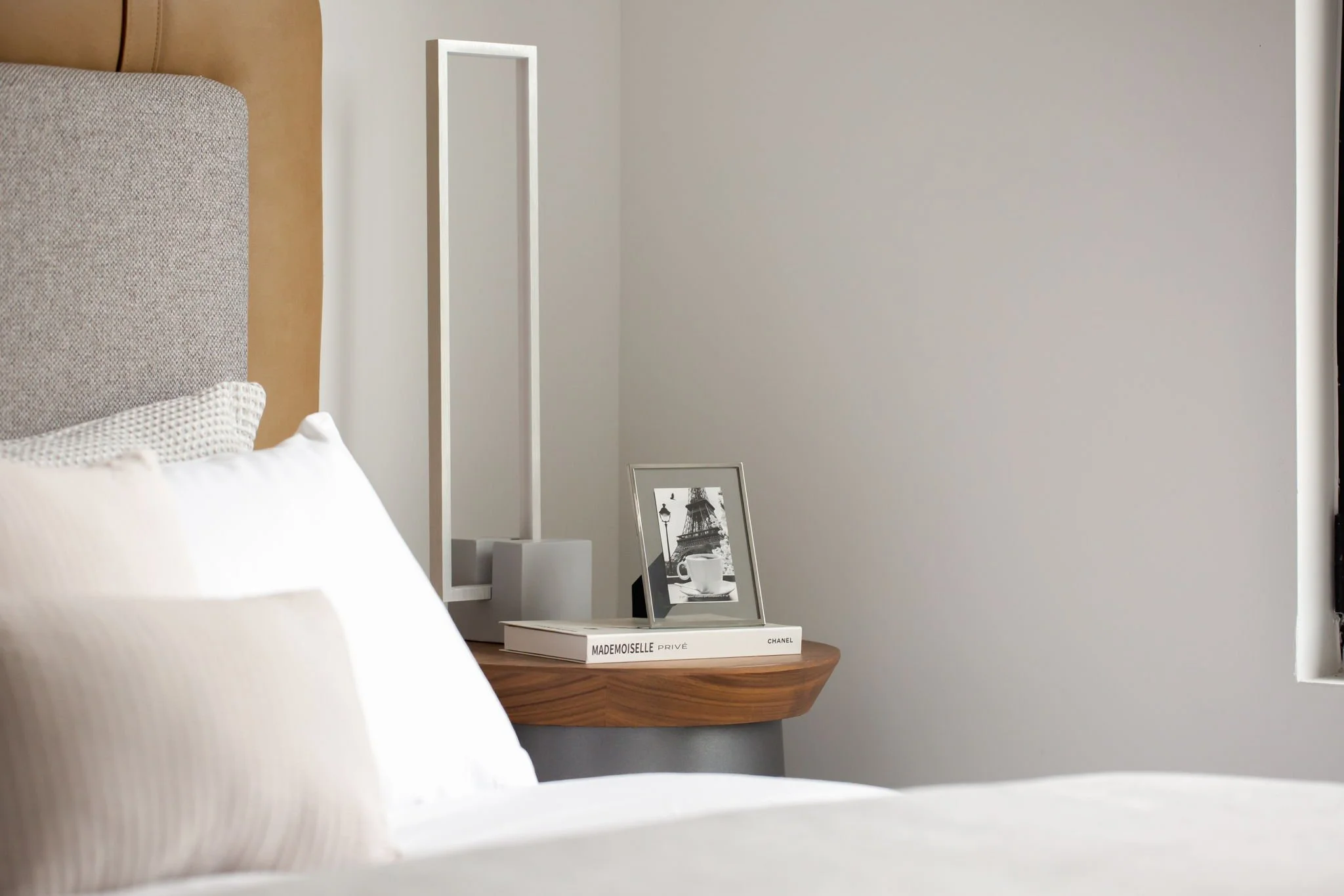 Close-up of a bedroom scene with a bed and a wooden nightstand. The bed has white linens and a textured pillow. On the nightstand, there are two books, a framed black-and-white photo, and a modern white lamp. The wall is white and part of a window is