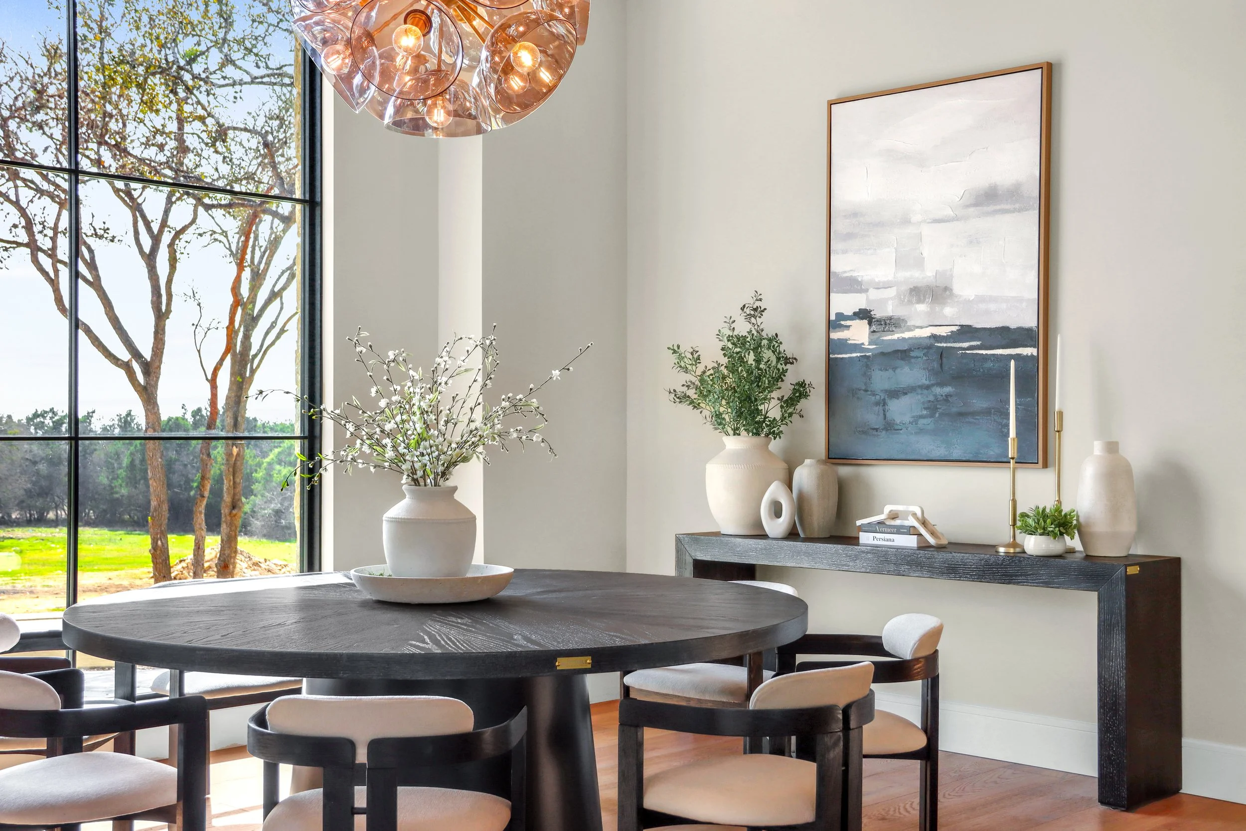 Modern dining room with a round black table, six white chairs, a vase with branches on the table, large window with a view of trees, abstract artwork on the wall, and a black sideboard with vases, books, and candles.