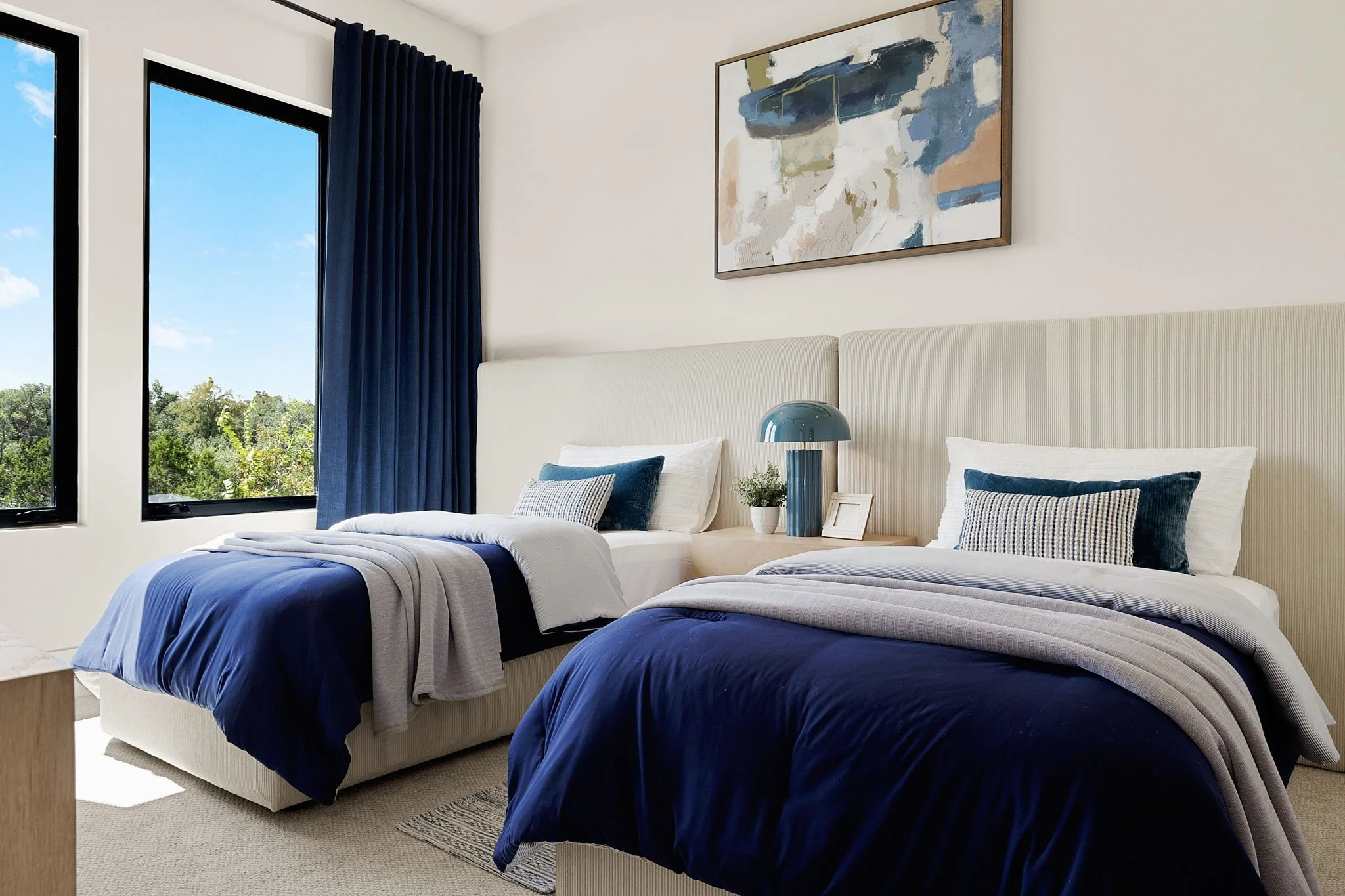 A bedroom with two single beds with blue and white bedding, dark blue curtains, and a large window showing green trees outside. There is a modern table lamp and artwork on the wall above the beds.