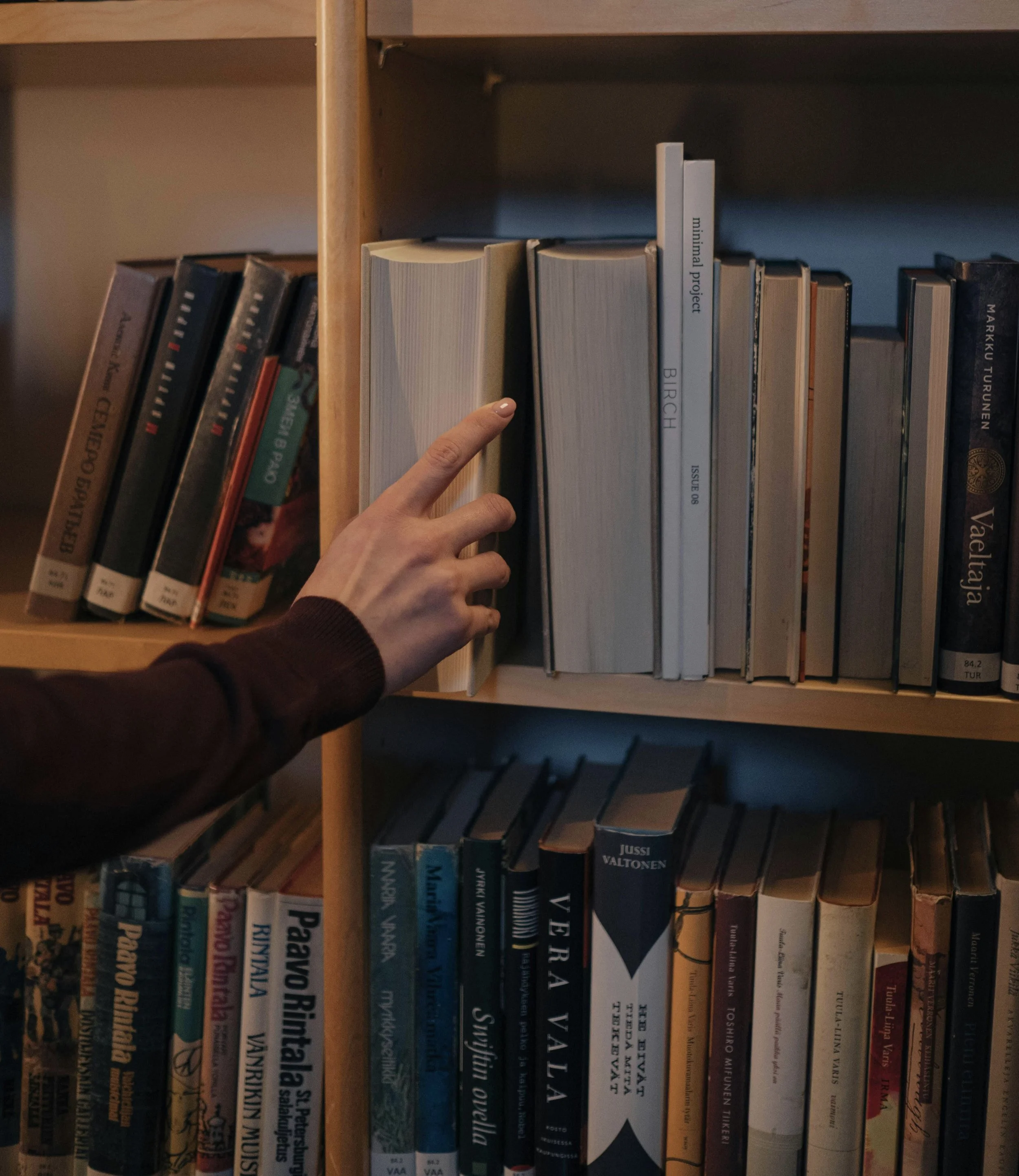 A person's hand reaching for a book on a wooden bookshelf, surrounded by books with various titles.