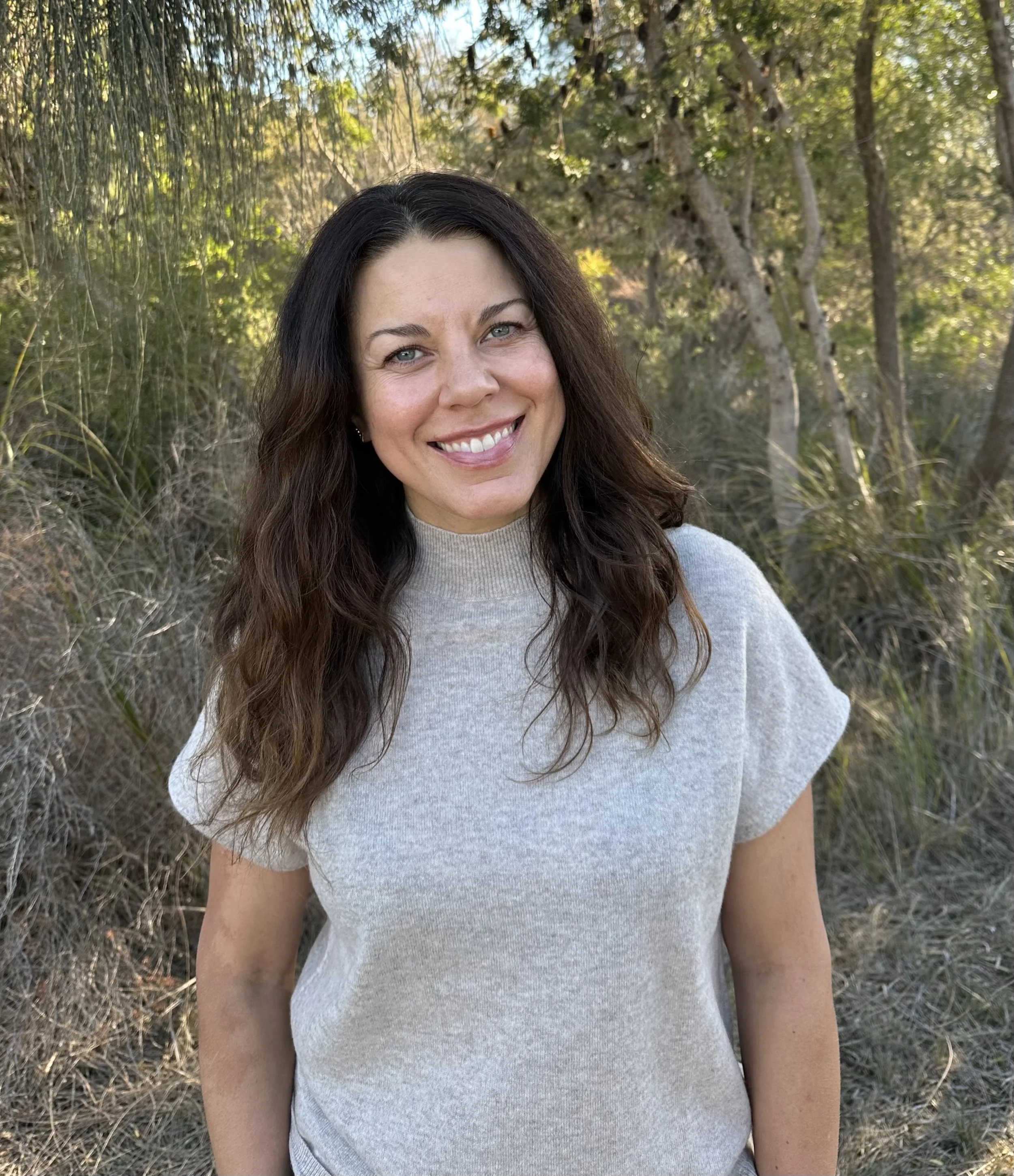 A woman with long dark brown hair and blue eyes smiling outdoors with trees and foliage in the background.