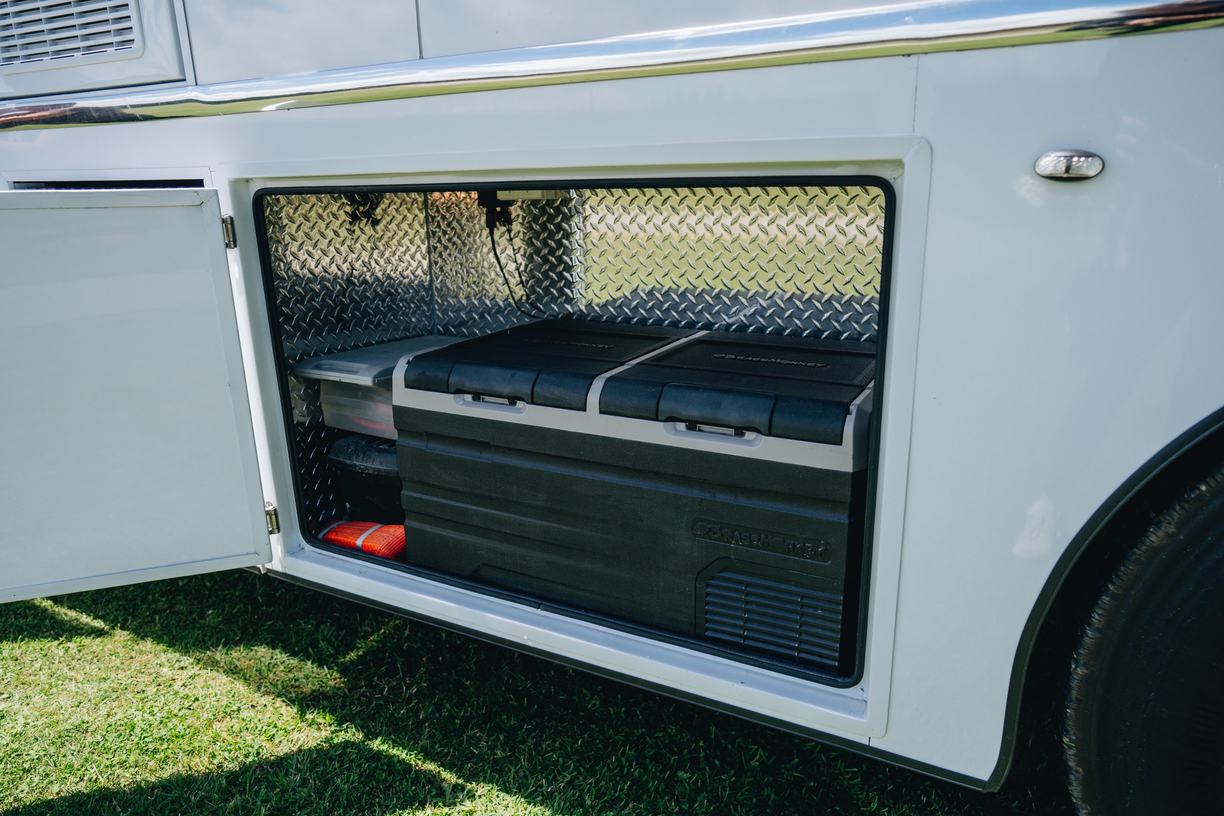 Open storage compartment on a white vehicle with diamond plate interior, containing a black and gray portable cooler and other equipment.
