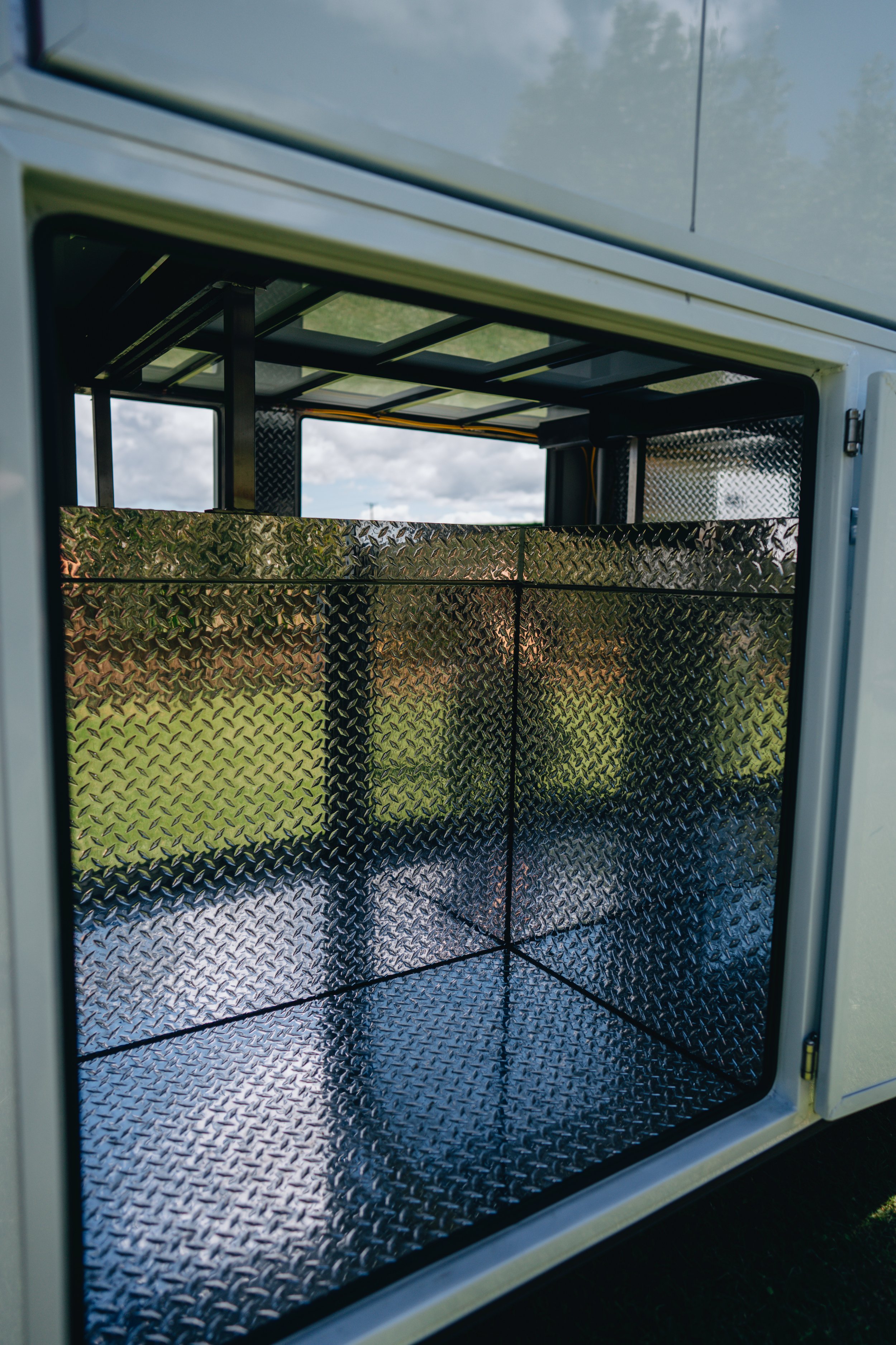 Open refrigerator with diamond plate interior panels and metal shelving, outdoors on grass and cloudy sky.