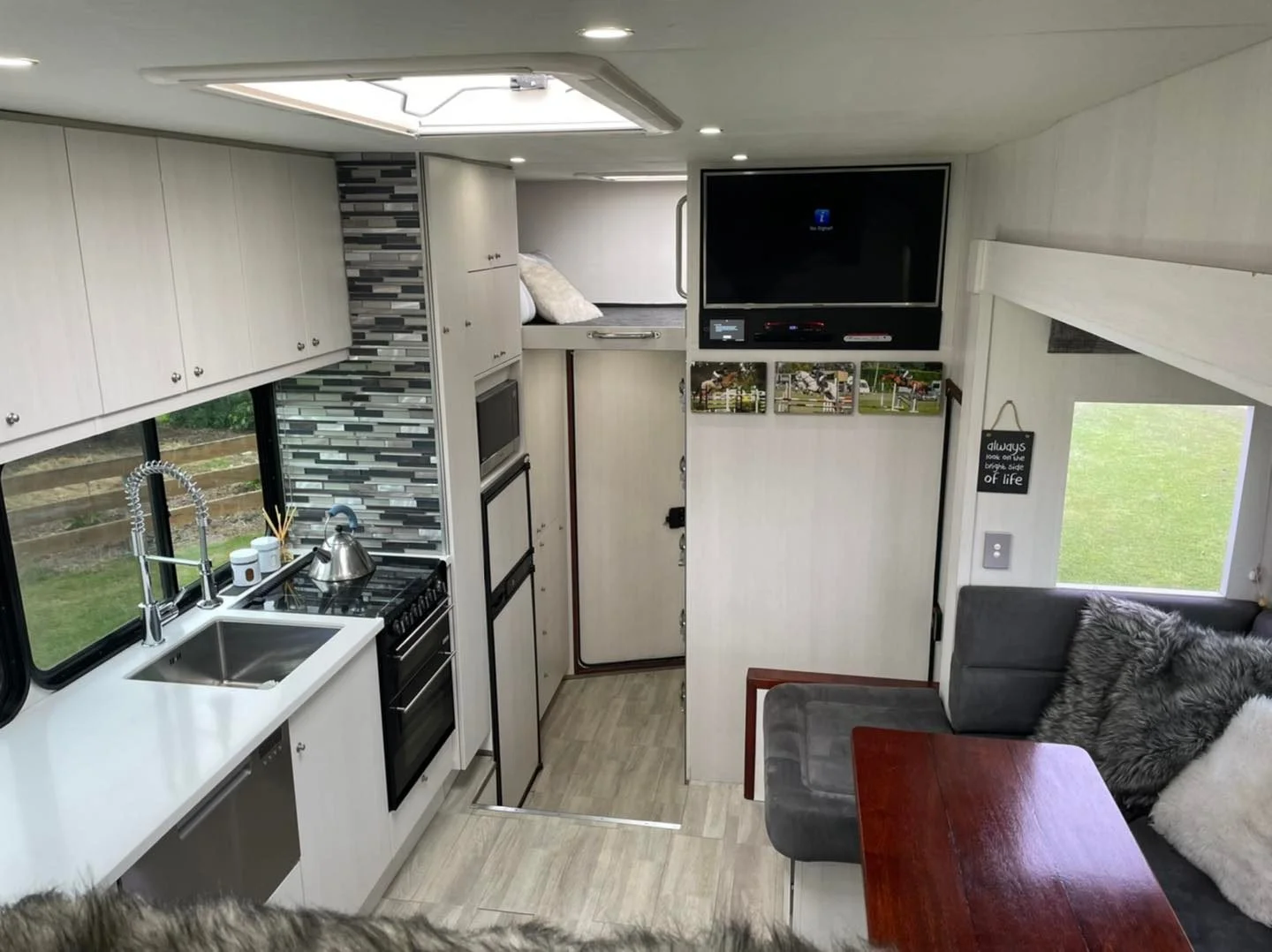 Interior of a compact camper or RV with a small kitchen, seating area, and a mounted television, featuring white cabinetry, a stainless steel sink, and a window with outdoor views.
