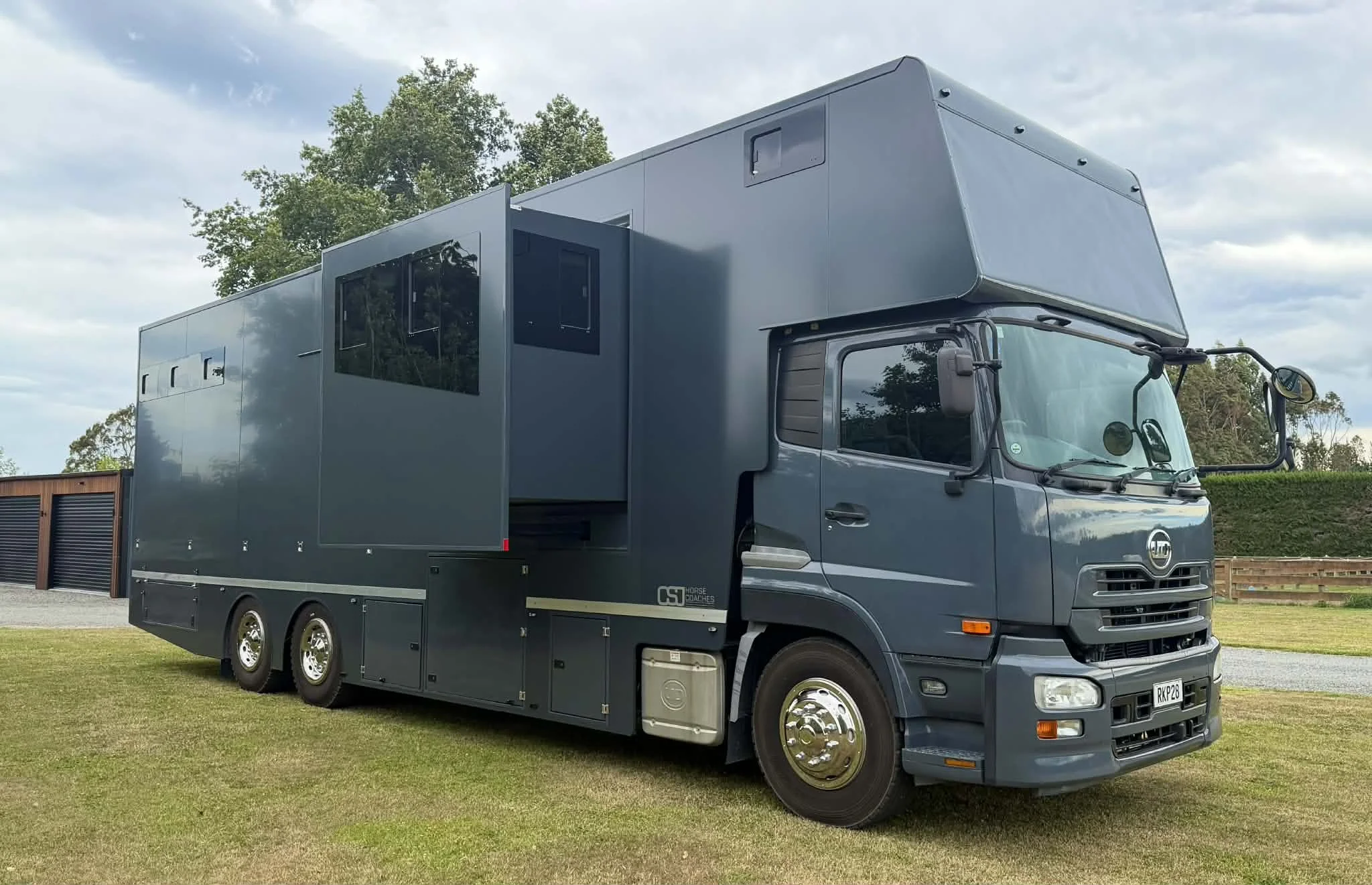 A large, custom-built, dark gray mobile command or living vehicle on a grassy field with trees and a partly cloudy sky in the background.