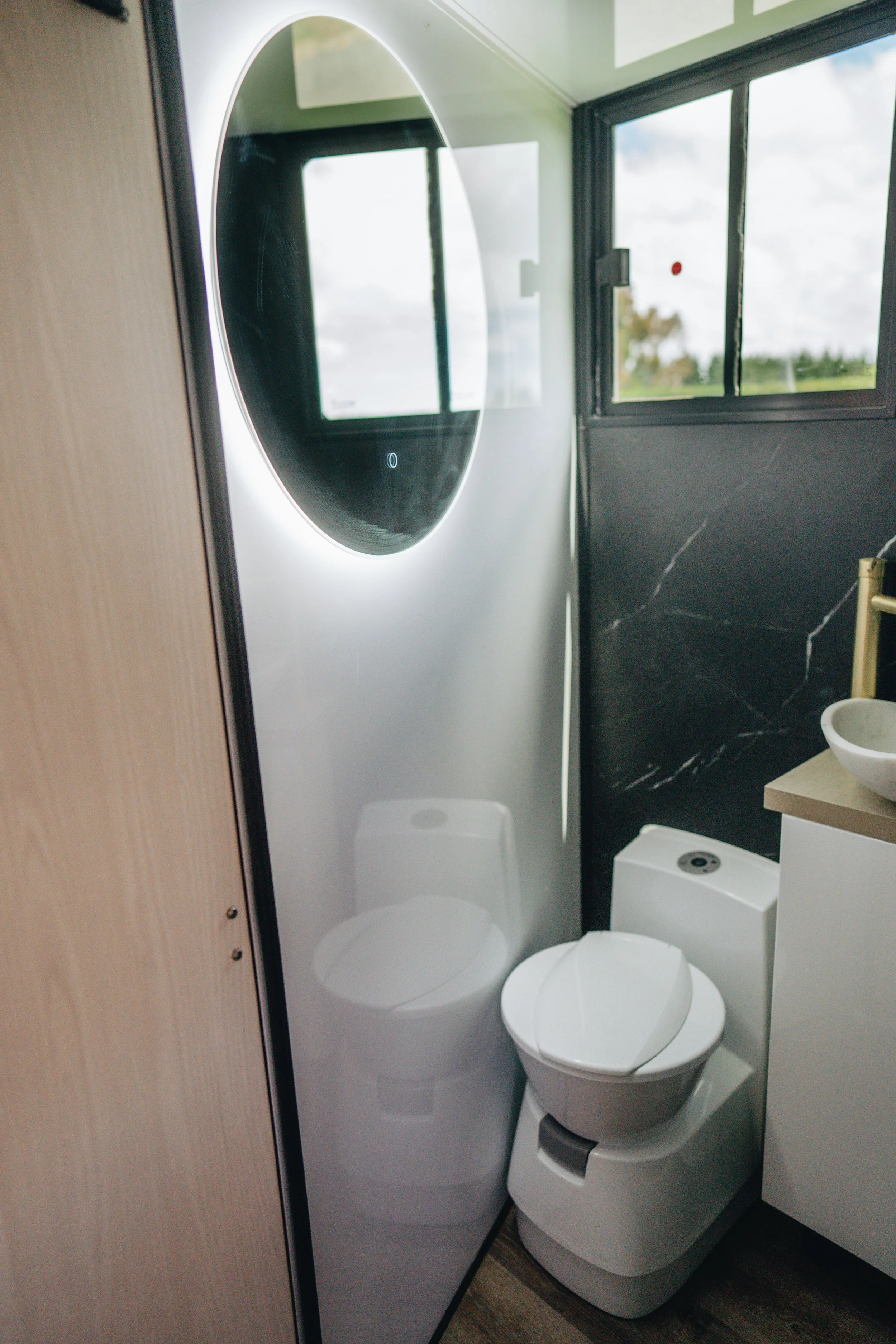 Modern bathroom with a wall-mounted mirror, a window, a toilet, and a small sink with a gold faucet.