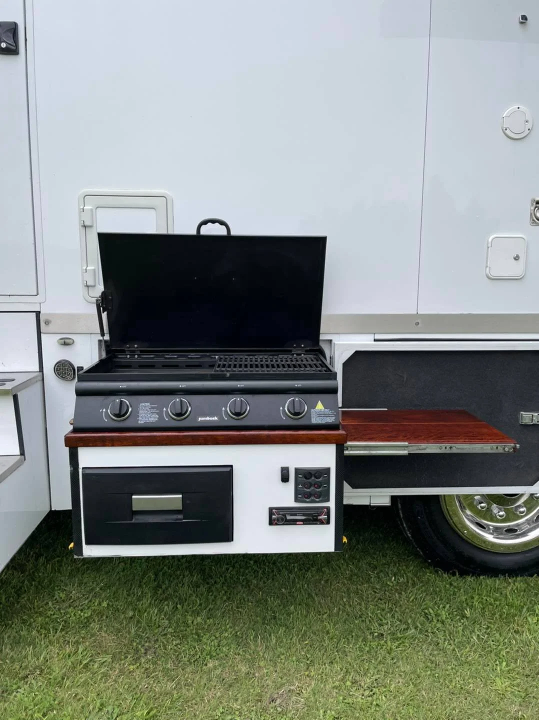 Portable outdoor kitchen with a black grill featuring four control knobs and a fold-down lid, set against a white mobile trailer with a small side table and wheels, on grass.