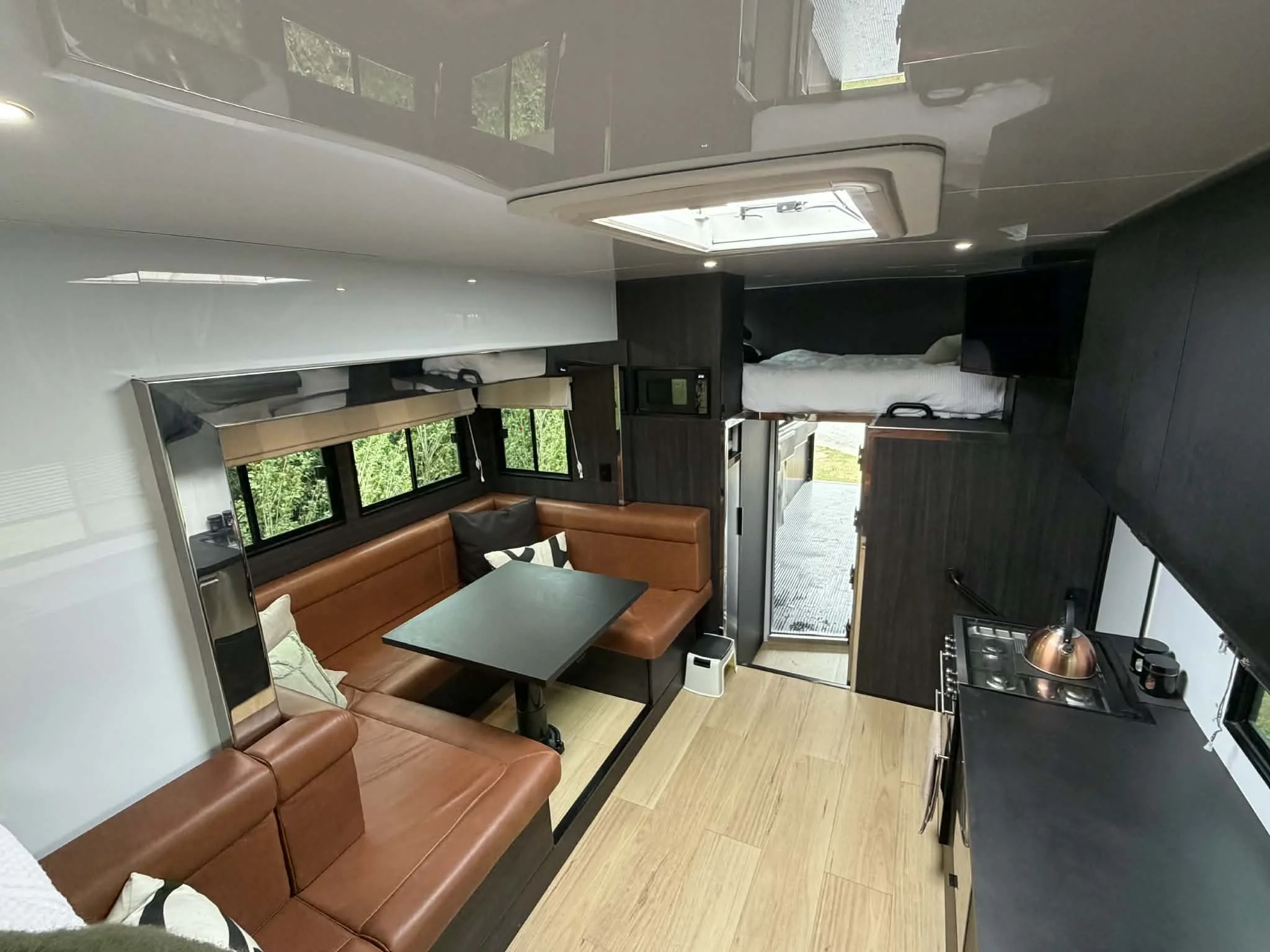 Interior of a modern camper or tiny home with a kitchen area, a dining booth with brown leather seats, a small bed platform, and windows showing greenery outside.