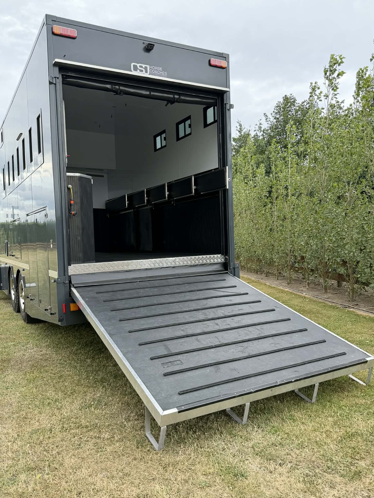 A black horse trailer with an extended ramp and interior windows, parked on grass.