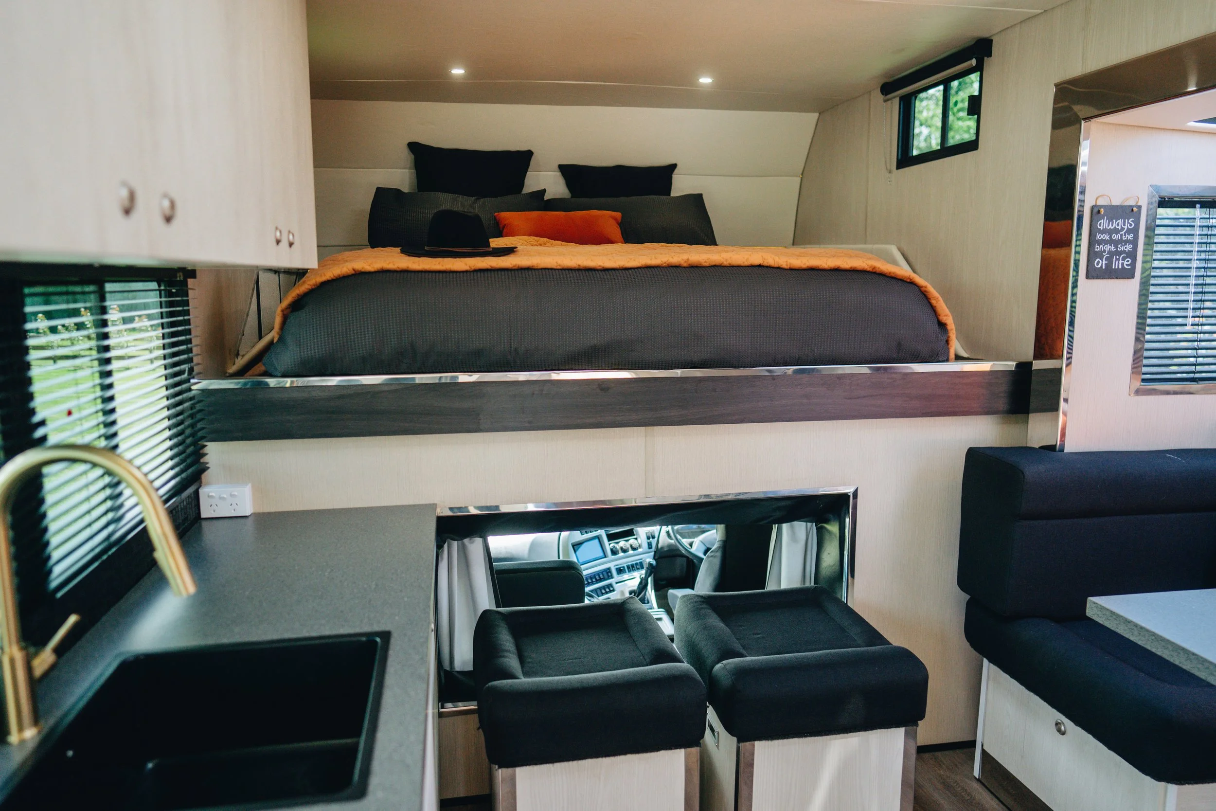Interior of a compact motorhome with a raised sleeping area, small kitchen with a sink, seating area, and a view of the driver's cockpit.