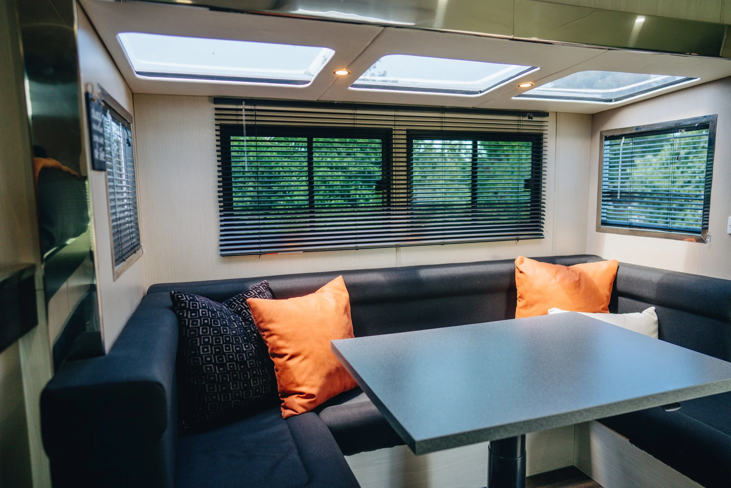 Interior of a living space with black L-shaped sofa, orange and patterned pillows, and windows with black blinds, with skylights on the ceiling.