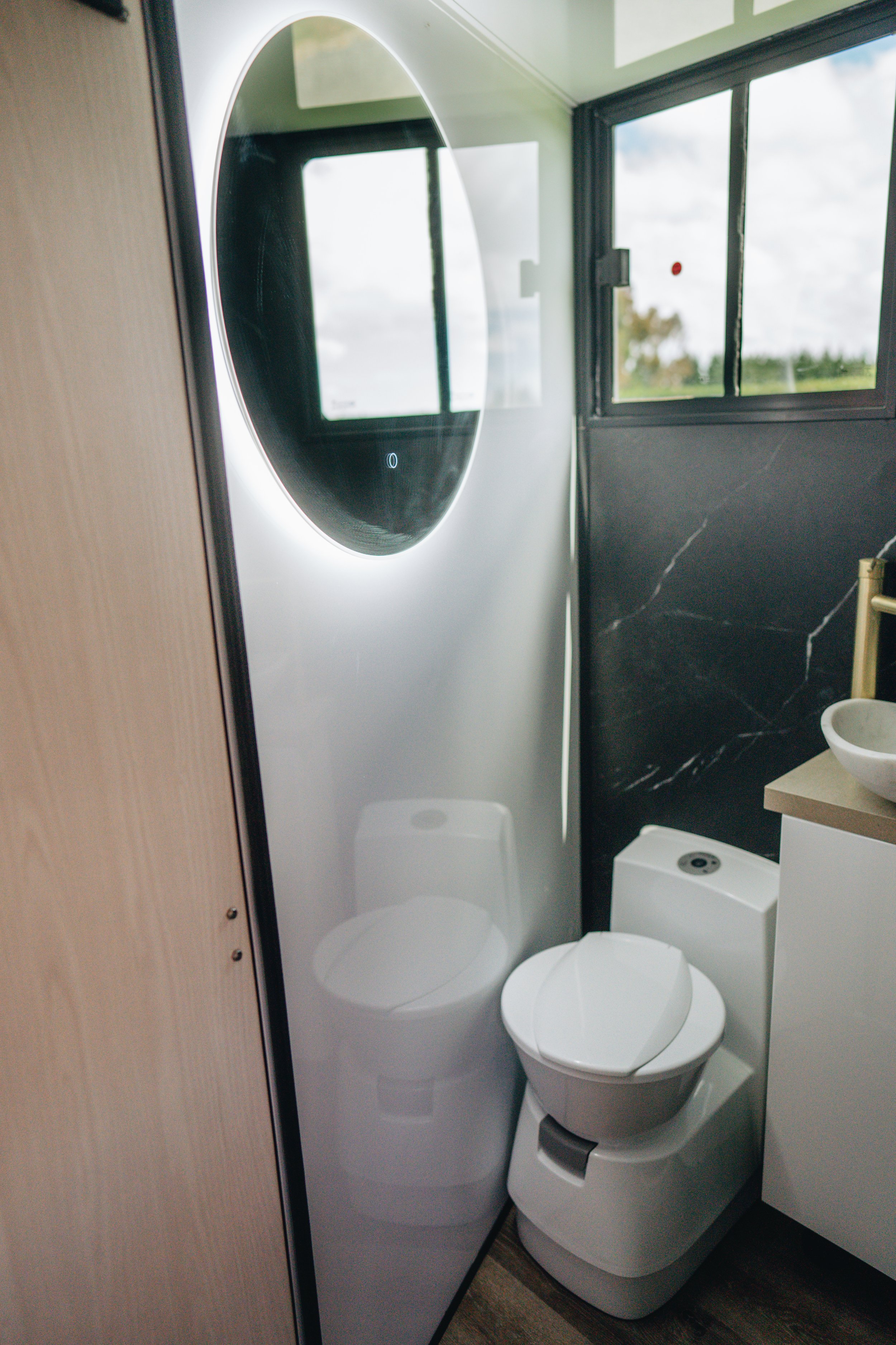 Modern small bathroom with a wall-mounted round mirror with LED lighting, a white toilet, a vanity with a vessel sink, and a black marble accent wall by a window showing an outdoor view.